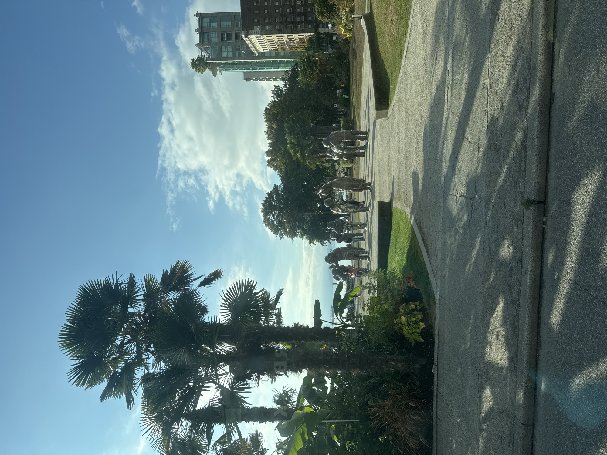 The photograph captures a sunny day in Vancouver, Canada, likely in the summer or early autumn, given the clear blue sky with scattered clouds. The scene is set in a park with lush greenery, featuring several prominent palm trees and other tropical-looking foliage on the left side of the frame. A paved pathway winds through the park, leading towards a group of metallic sculptures in the middle ground. These sculptures depict figures that appear to be engaged in some form of activity, possibly sports or a communal gathering, with several of them looking towards the viewer. The figures are life-sized and have a somewhat abstract, metallic finish.
In the background, several buildings rise above the trees. To the right, a brick building with numerous windows is visible, and to its left, a modern, taller building with a distinctive green glass exterior and a unique top featuring trees and what appears to be a helipad or observation deck. The presence of these buildings suggests an urban setting adjacent to the park.
Beyond the park and the sculptures, a body of water can be seen in the distance, with a large cargo ship sailing on it. This indicates the park is located near the waterfront. The pathway itself is made of concrete slabs and shows some signs of wear and tear, with cracks and discoloration. Sunlight casts dappled shadows on the pathway, indicating a bright, sunny day. A few individuals, including a child, are visible near the sculptures, seemingly observing them or passing by. The overall impression is of a pleasant public space within an urban environment, showcasing art and natural elements with views of the city and the water.