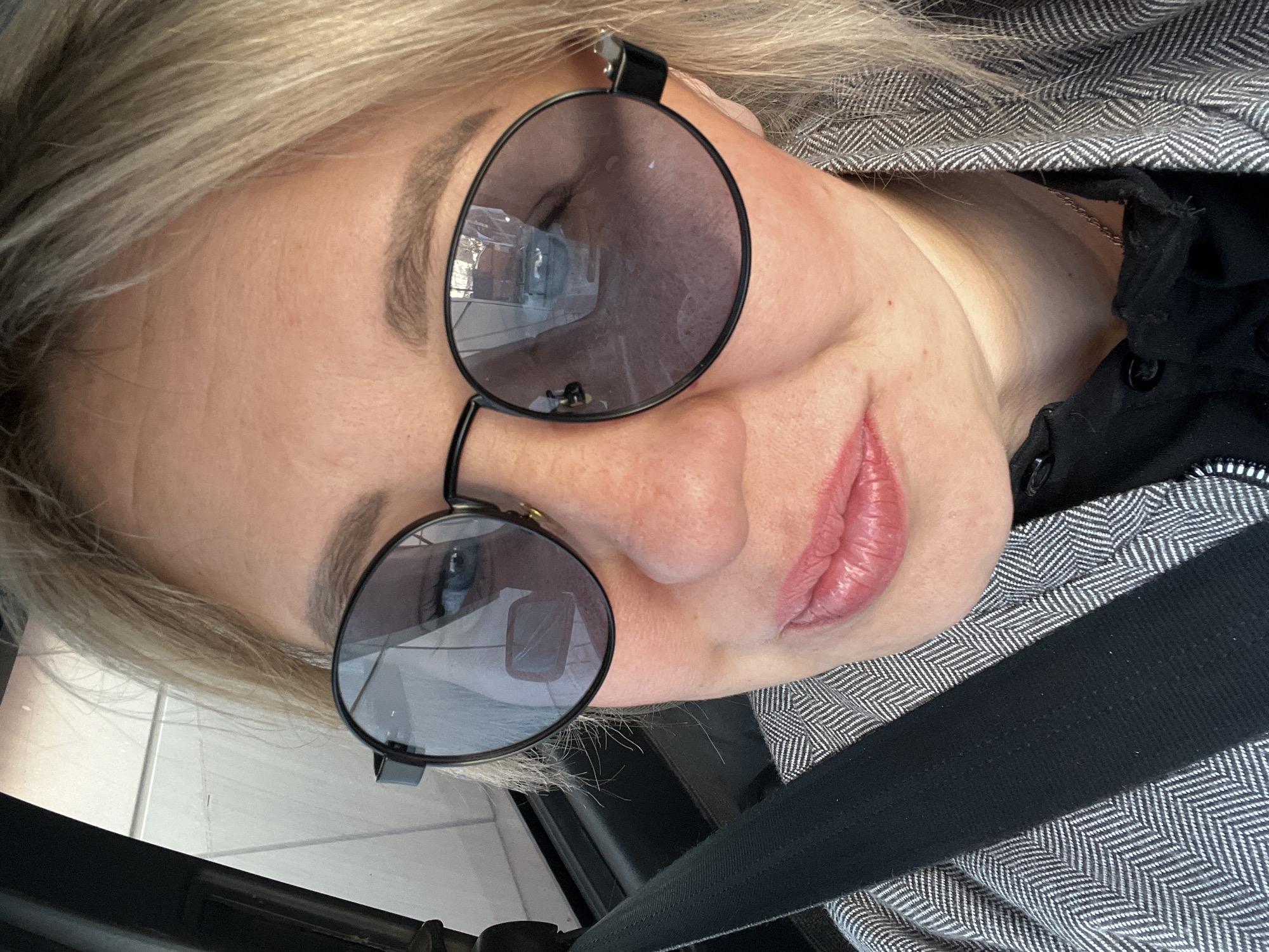 A close-up selfie captures a light-haired woman, angled as if looking upwards, likely inside a vehicle in Chaikovsky, Russia. She wears stylish round, dark-lensed sunglasses with thin black frames. Reflections in the sunglass lenses show a bright, possibly indoor or structured environment. Her left eyebrow, with neatly defined dark hairs, is visible above the glasses, and her skin appears clear. Her lips are slightly parted, revealing a soft, natural pink color, and she has a subtle, relaxed smile.
She is dressed in a black top or shirt, and a distinctive grey and white herringbone patterned jacket or coat. A thin metallic chain necklace is partially visible around her neck, tucked beneath her jacket. A black seatbelt crosses diagonally from the bottom right of the frame, confirming she is secured within a vehicle. The background behind her head appears to be a light-colored, possibly paneled or tiled, surface. No text is visible in the image. The overall impression is a casual self-portrait taken while in transit.