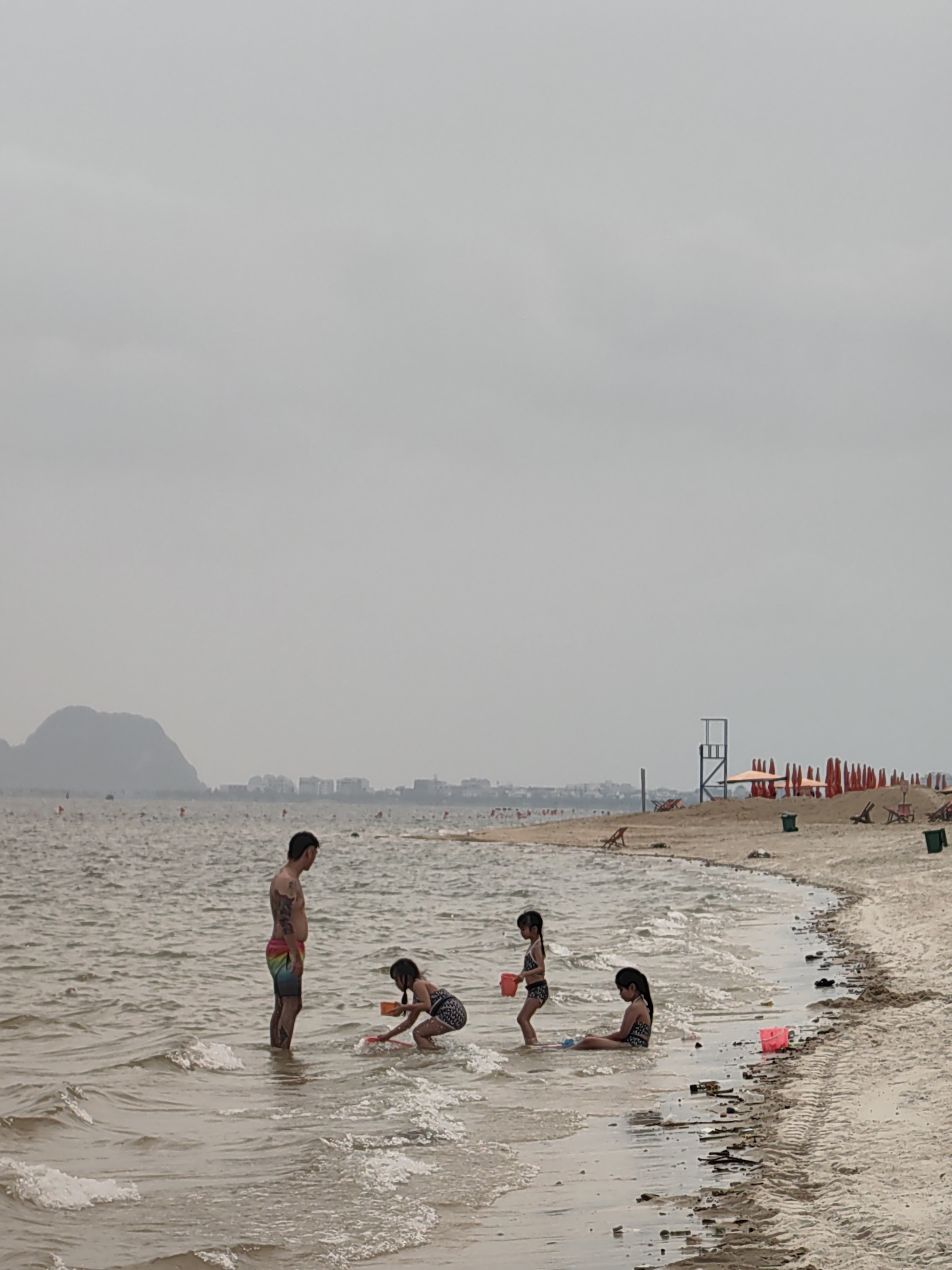 On a cloudy day in Bai Chay Ward, Vietnam, a man and three children are playing in the shallow water at the beach. The man is standing in the water while the children are playing with sand toys. In the background, there is a lifeguard tower and a row of beach umbrellas and chairs on the sand. There are buildings and a mountain in the far background.