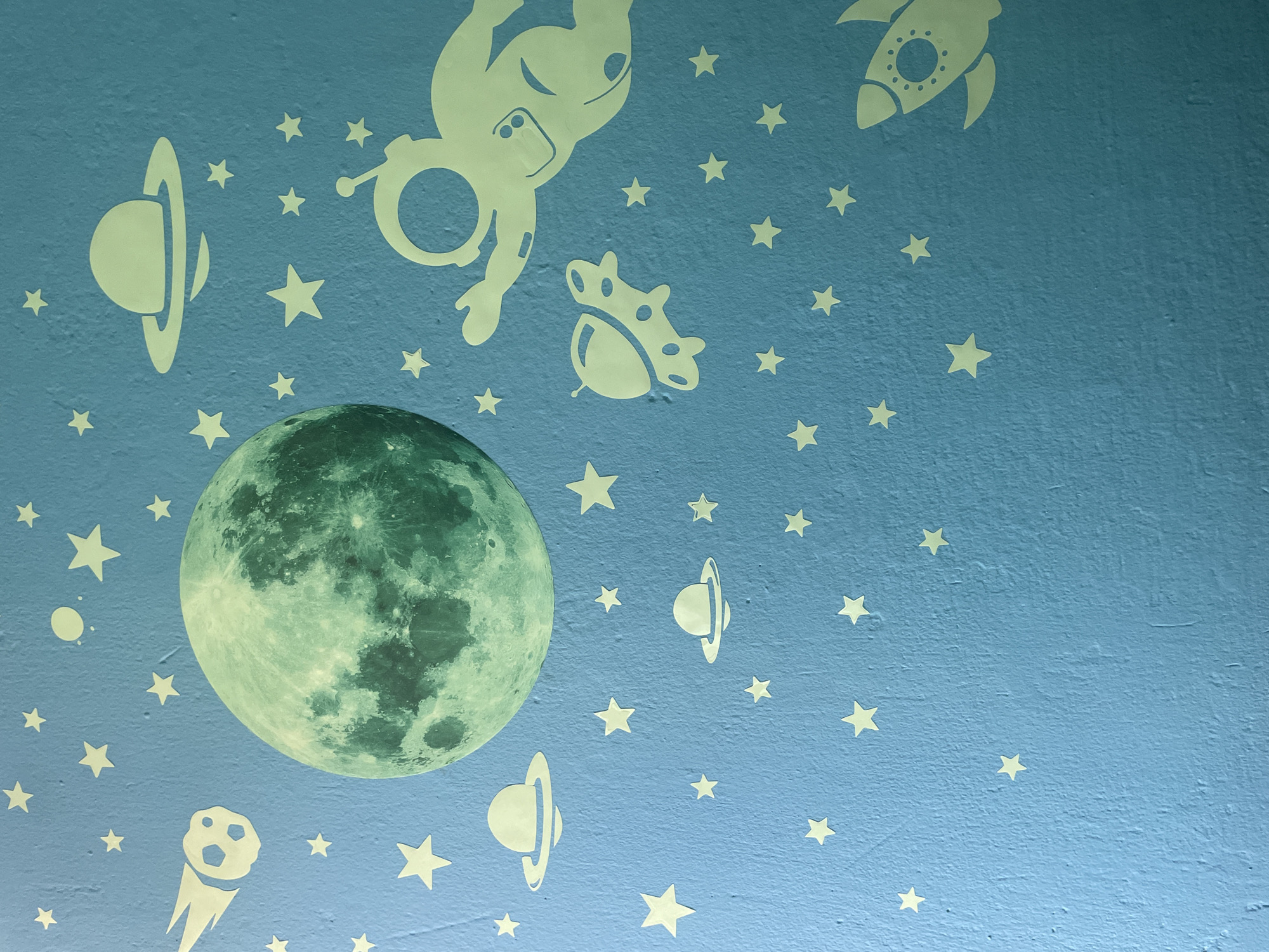This is a photograph of a blue wall decorated with glow-in-the-dark stickers depicting a space theme. The main subject is a large, realistic-looking moon positioned on the left side of the frame. Surrounding the moon are various stylized celestial objects and figures. These include several five-pointed stars scattered across the wall, a rocket ship in the upper right corner, an astronaut floating in the upper left, and two ringed planets, resembling Saturn. Additionally, there are depictions of flying saucers and a comet with a trailing tail. The overall scene conveys a playful and imaginative atmosphere, suggesting a child's room or a themed decoration. The lighting in the image indicates it was taken in a room, likely at night, given the glow-in-the-dark elements. There is no visible text in the image. The location context provided is Kołobrzeg, Poland, but this information cannot be visually verified from the image itself.