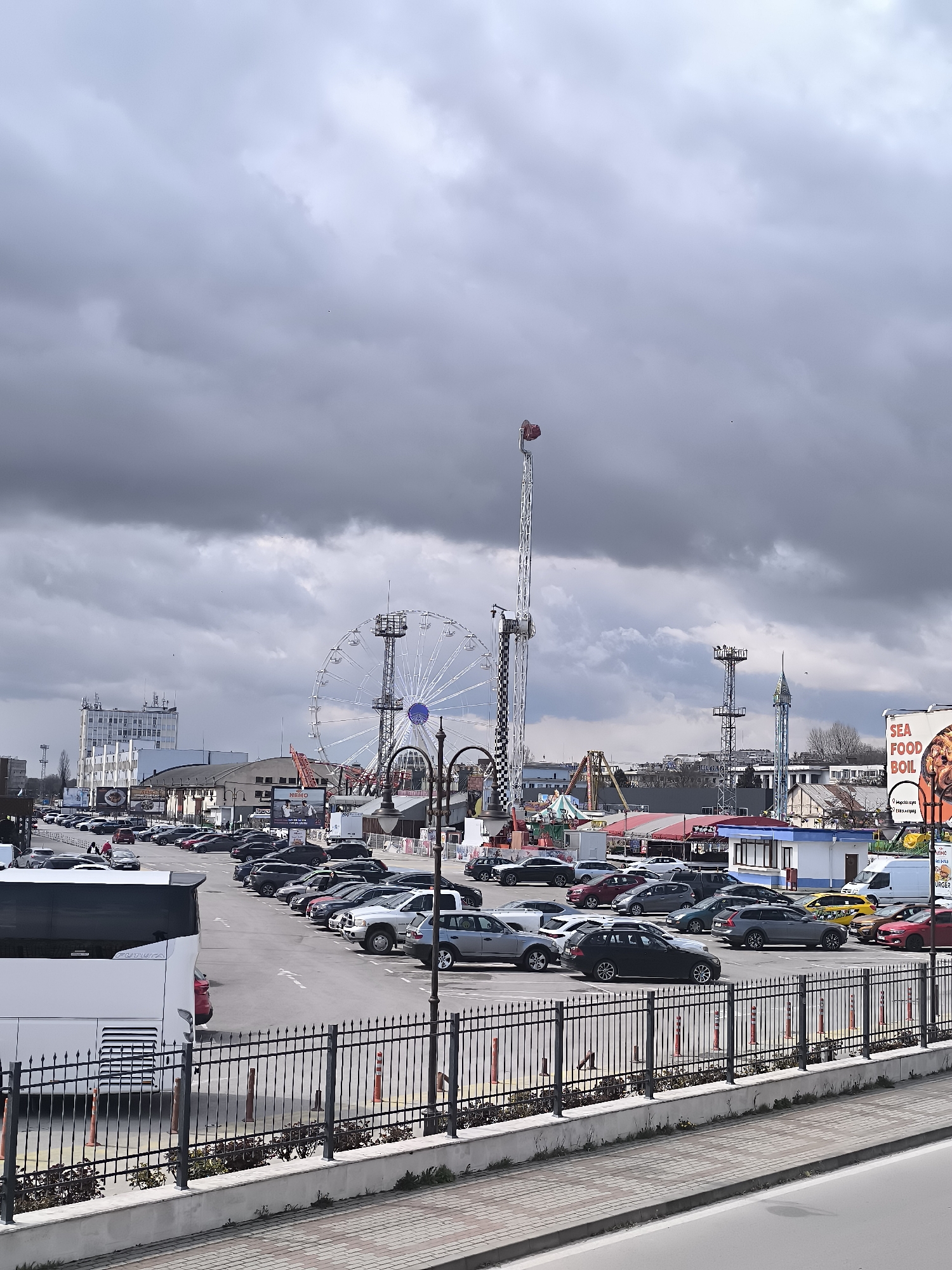 The image depicts a parking lot adjacent to an amusement park under a cloudy sky. Numerous cars are parked in an organized fashion within the lot, bordered by a black iron fence along the foreground. Behind the parking lot, the amusement park is visible, including a Ferris wheel and a tall drop tower. In the background, buildings and structures can be discerned, including a larger white building, suggesting an urban or suburban location. The image suggests the location is in Varna, Bulgaria due to the presence of the amusement park and the architecture visible.