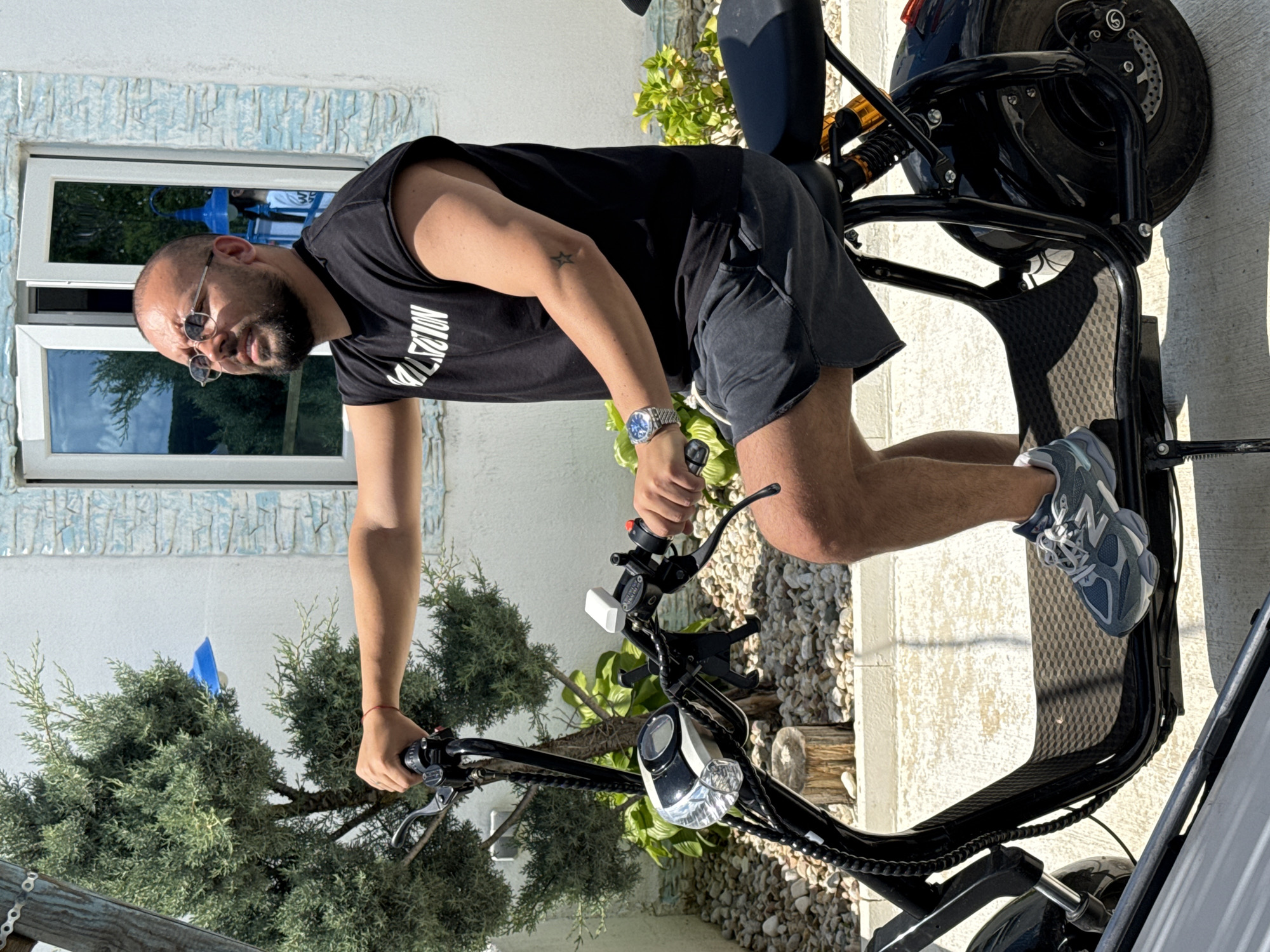 In Baliovtsi, Bulgaria, a man with a beard and glasses, wearing a black t-shirt with the word "VALUATION" in white lettering and dark gray shorts, sits on a black electric scooter. He has a watch on his left wrist and a small star tattoo on his right bicep. The scooter has a digital display on the handlebars and a reflective headlight. The man is holding the handlebars, with his left hand on the brake lever. He is looking to his right. The background features a white building with blue trim around the windows, a small evergreen tree, and a ground covered in small pebbles. The scene appears to be outdoors on a sunny day.