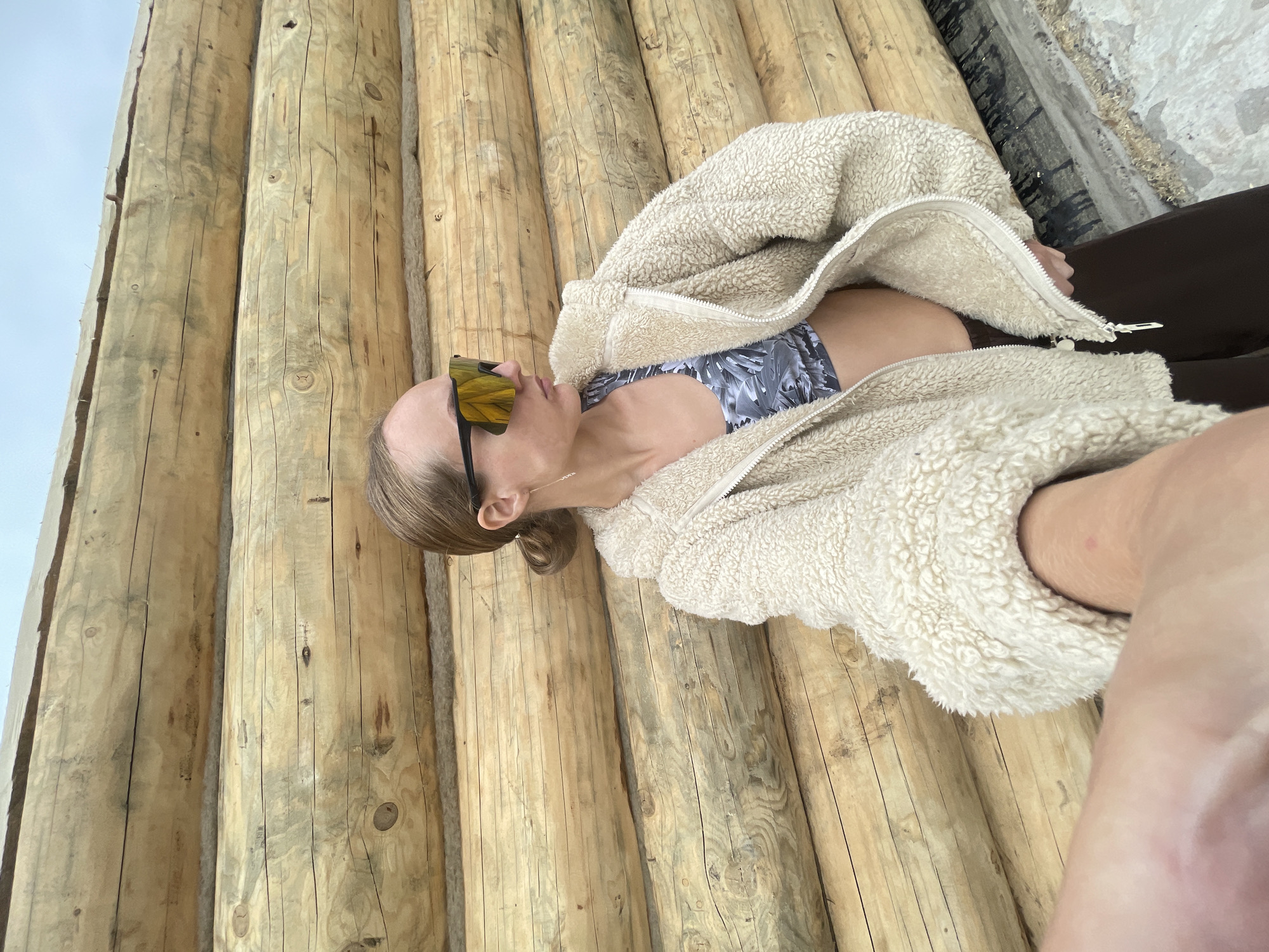 A woman in sunglasses, a patterned sports bra, a fuzzy cream-colored jacket, and brown pants poses for a selfie in front of a wall made of stacked, unfinished logs. The wall is oriented horizontally, and the logs have visible knots and natural imperfections. The woman's left arm and hand are visible in the foreground, holding what appears to be a phone to take the picture. Her hair is pulled back in a low ponytail, and she is wearing long, thin earrings. The ground beneath her is a mix of sand and what looks like wood shavings. The image appears to have been taken outdoors, possibly in a rural or construction setting. There are no other people visible in the frame. The sky is overcast and visible at the top of the image. The location is identified as Podshivalovo, Russia.