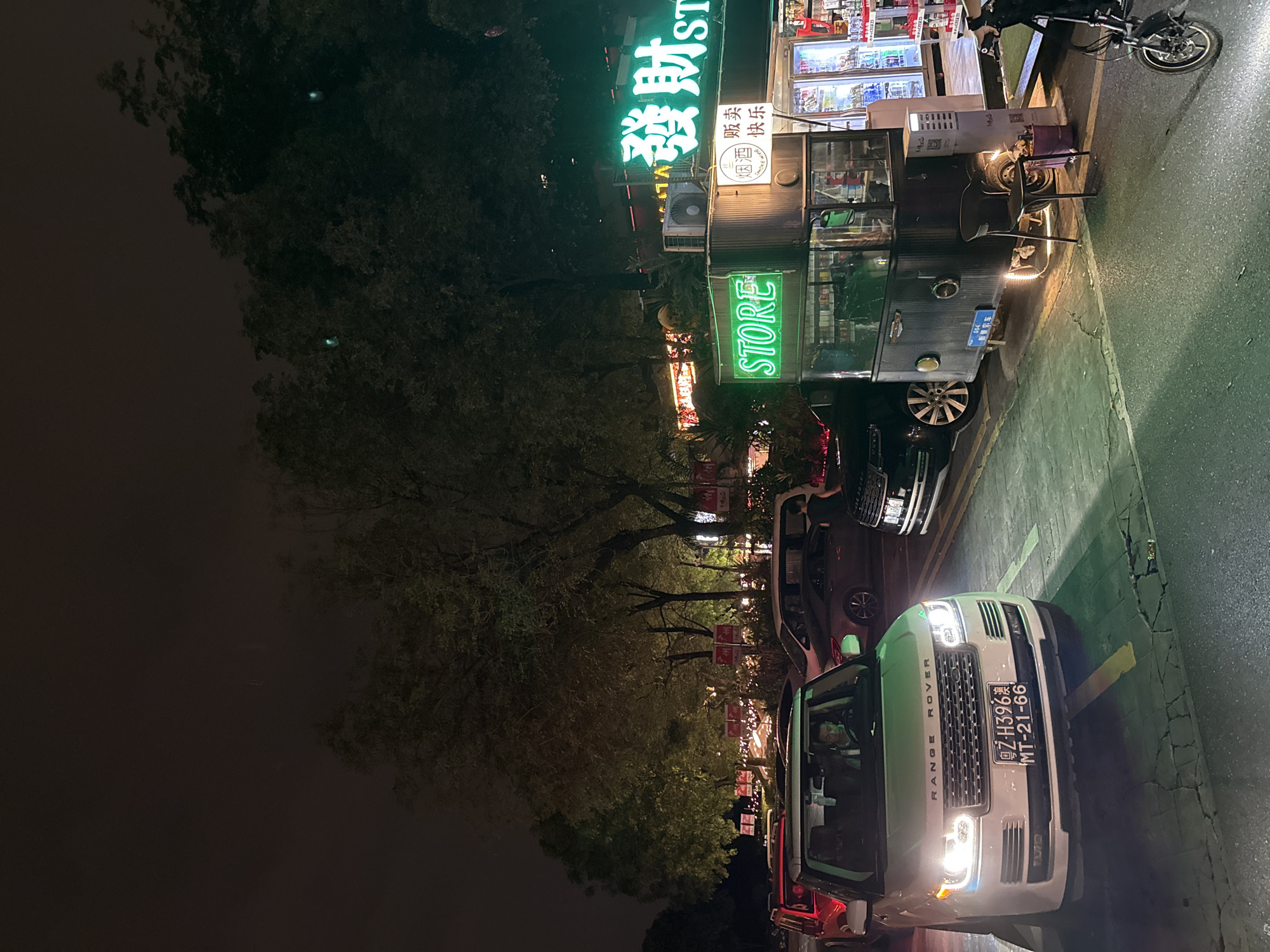 Here's a detailed description of the image:
**Overall Scene:** The image shows a nighttime street scene in what appears to be a commercial district. The image is oriented sideways. There are cars parked along the curb, trees lining the street, and businesses visible with illuminated signs.
**Specific Details:**
* **Vehicle:** A light-colored "Range Rover" car is parked prominently in the foreground. The license plate is visible, reading "Z-H396澳 MT-21-66" (likely a Macanese license plate).
* **Street:** The street is paved, with visible cracks and repairs.
* **Businesses:** There's a "STORE" visible in what looks like a converted bus. The signs on the store are in Chinese, including characters that translate to "Prosperous Fortune ST". The store sells beer and has a liquor license.
* **Other parked cars:** There are multiple parked cars beside the Range Rover.
* **Street Lighting:** The street is well-lit with streetlights and the illumination from the businesses.
* **Trees:** Trees line the street, with their foliage partially obscuring some of the buildings and sky.
* **Atmosphere:** It seems to be a calm night.
**Inferences about Location:**
* Based on the language on the signs, location of store in a bus, and the license plate of the car, the photo was taken in China. The License plate has a location identification for the city of Zhuhai in the Guandong province next to Macau, which is in turn next to Hong Kong. Thus, the image was likely taken in the Tianhe district of Guangzhou, Guangdong, China.