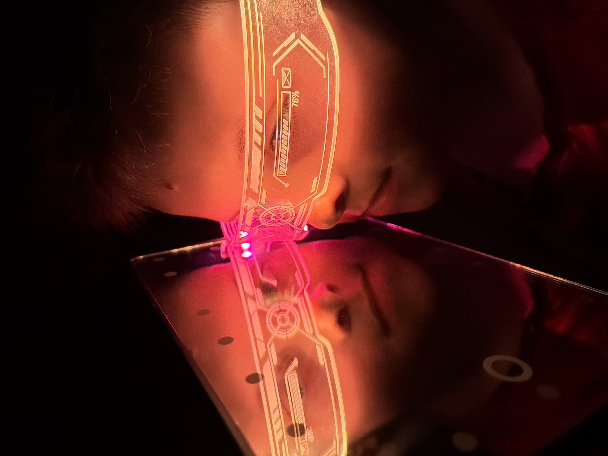A person lies prone, their face illuminated by the pinkish-red glow of futuristic-looking, possibly digital glasses or a visor. The glasses display a white, digital-style graphic including a battery-like bar showing 76%. A reflective surface below the person's face mirrors the light and the image of the glasses. The surrounding environment is dark, with minimal detail visible. The image location is not discernible based on the provided visual information.