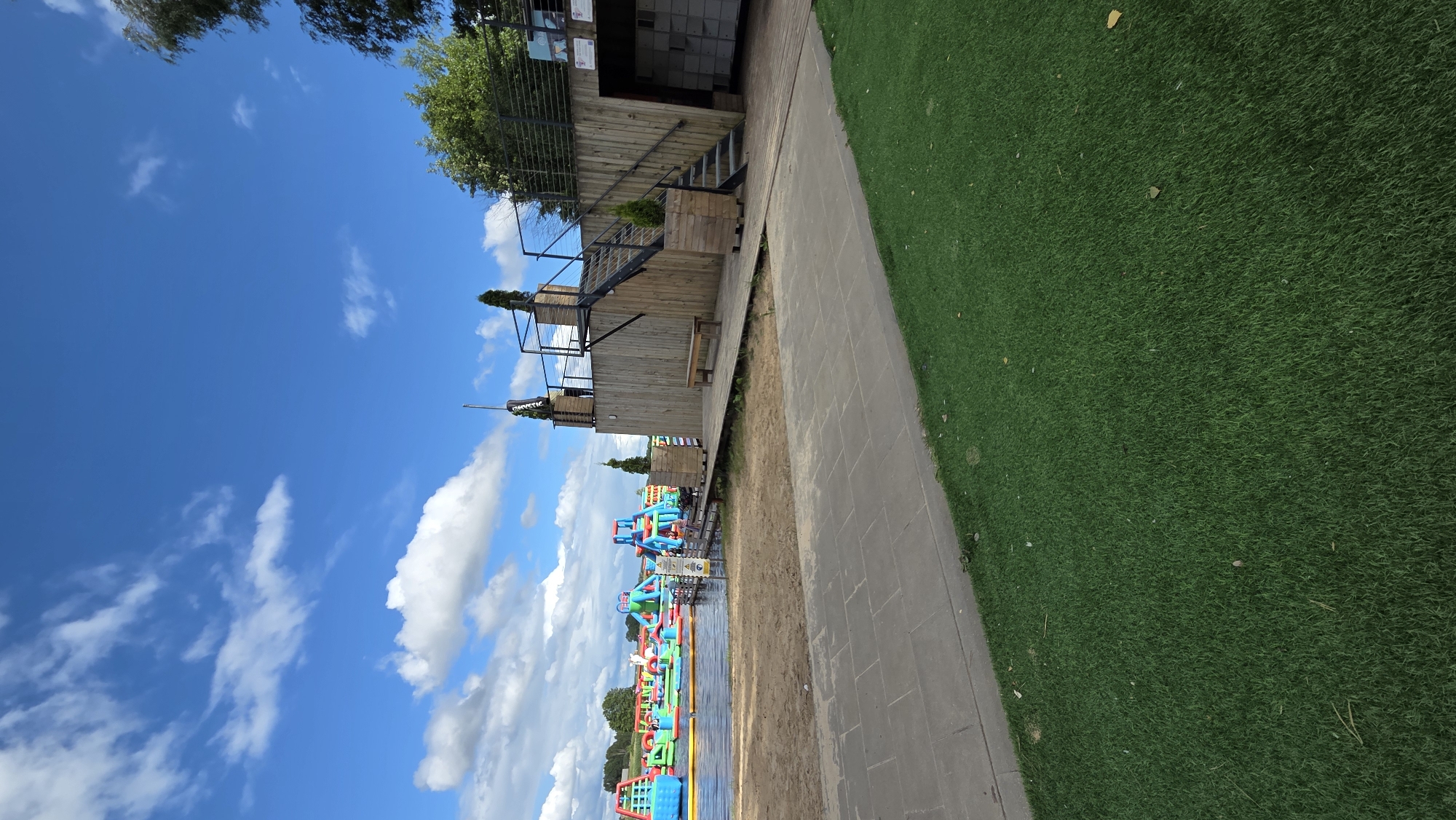 An outdoor scene features a large inflatable water park on a lake in the background, with multiple colorful slides and obstacles. In the foreground, there is a sandy area transitioning to a paved walkway and then an area of artificial turf. To the right, a wooden structure with stairs leads up to a platform. There is a bench on the wooden platform. The sky is blue with scattered white clouds. The location is identified as being in Jaunmārupe, Latvia.