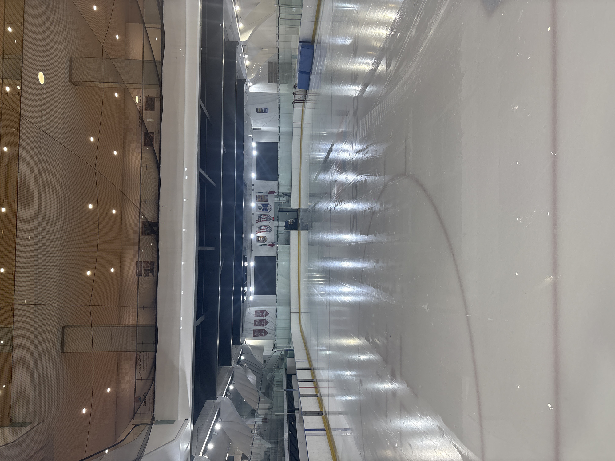 The image depicts the interior of an ice hockey rink. The ice surface is pristine, with visible markings and lines typical of a hockey rink. A net on the left of the image appears to be preventing any potential pucks from hitting the surrounding crowd. Benches are present on the far side of the rink, which is surrounded by a dark wall that appears to overlook the ice surface from a higher vantage point. A series of banners and displays are visible along the wall, but the writing is indiscernible. The overhead lighting is bright and reflects off the ice, creating a stark contrast. Based on the architectural style and promotional elements visible, the location may be the Ice Dome in Mexico City, Mexico.