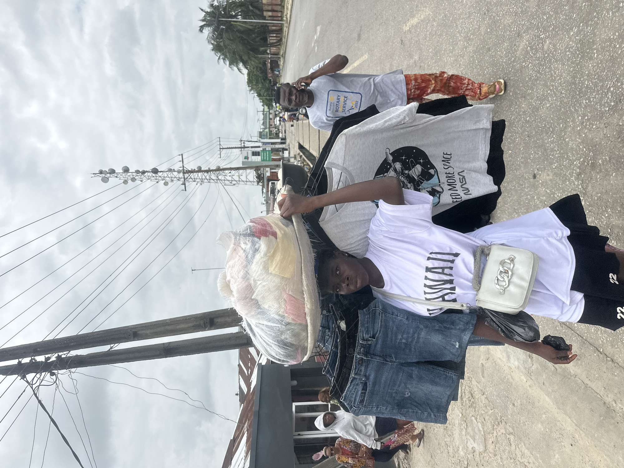 A young woman is pictured carrying a large bundle of clothes on her head and a rack of jeans and t-shirts on her shoulder. She is walking on a paved road in Badagry, Nigeria. The sky is overcast, and there are utility poles and a cell tower in the background. Other people are walking on the side of the road. A man in a grey t-shirt with "Rotary Service" printed on it is seen talking on his phone in the background. The scene appears to be a busy street market or a commercial area.