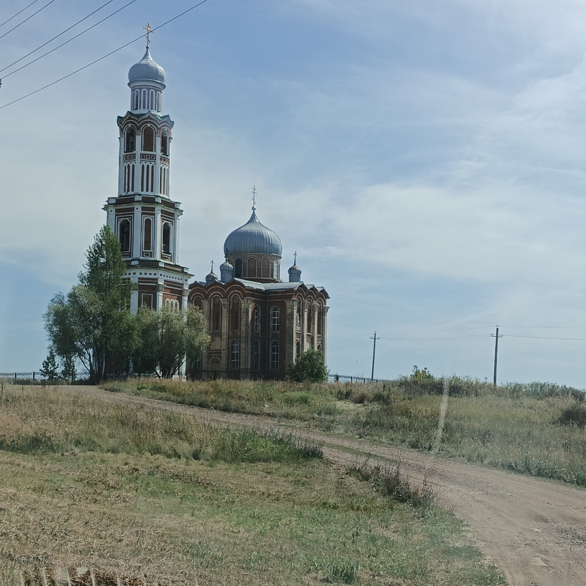 A full-shot photograph captures a red-brick church with a blue dome and a tall, multi-tiered bell tower against a pale blue sky with scattered white clouds. The church is situated on a grassy, slightly sloped hill in the village of Tugustemir, Russia. A dirt road leads towards the church from the foreground. The area surrounding the church is covered in dry grass and scattered green vegetation, with a few trees visible to the left of the bell tower. Power lines run across the top of the image. The scene depicts a quiet, rural landscape under daylight.