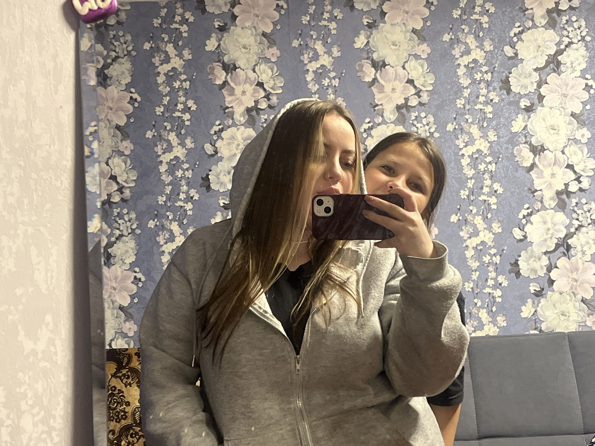 Two females pose for a selfie using a smartphone in what appears to be a residential interior. The primary subject, partially obscured by her own phone, wears a gray hooded zip-up sweatshirt, with her hood up. Another female is seen behind her, partially visible. The background features a floral patterned wallpaper. Elements of furniture such as a couch and part of a wall decoration are present. Location in Novopokrovskaya, Russia is unconfirmed without further information.