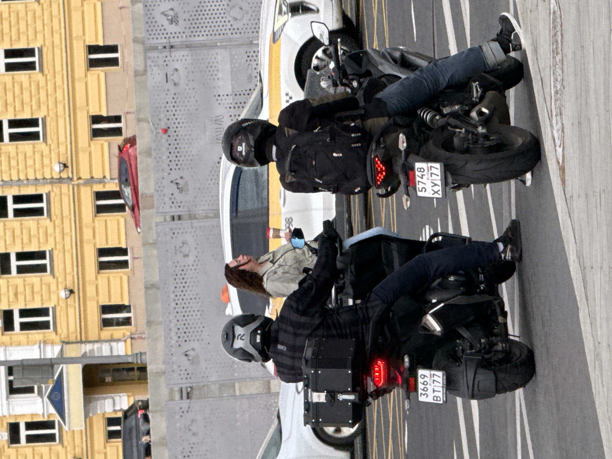 Two individuals on motorcycles are stopped at a street crossing in Moscow, Russia. The motorcycle on the left is ridden by a person wearing a black plaid jacket and a grey and black helmet. The motorcycle has a large black cargo box on the rear and a license plate reading "3669 BT 77". The motorcycle on the right is ridden by a person wearing a black jacket and a black helmet. This motorcycle has a black backpack and a license plate reading "5748 XY 77". A woman with long brown hair, wearing a light-colored jacket, is standing near the motorcycle on the left, holding a cup. A white taxi with yellow accents is visible in the background. The scene takes place on an asphalt road with lane markings. Buildings with yellow facades and white trim are visible behind a grey decorative fence. A red car is also visible in the distance.