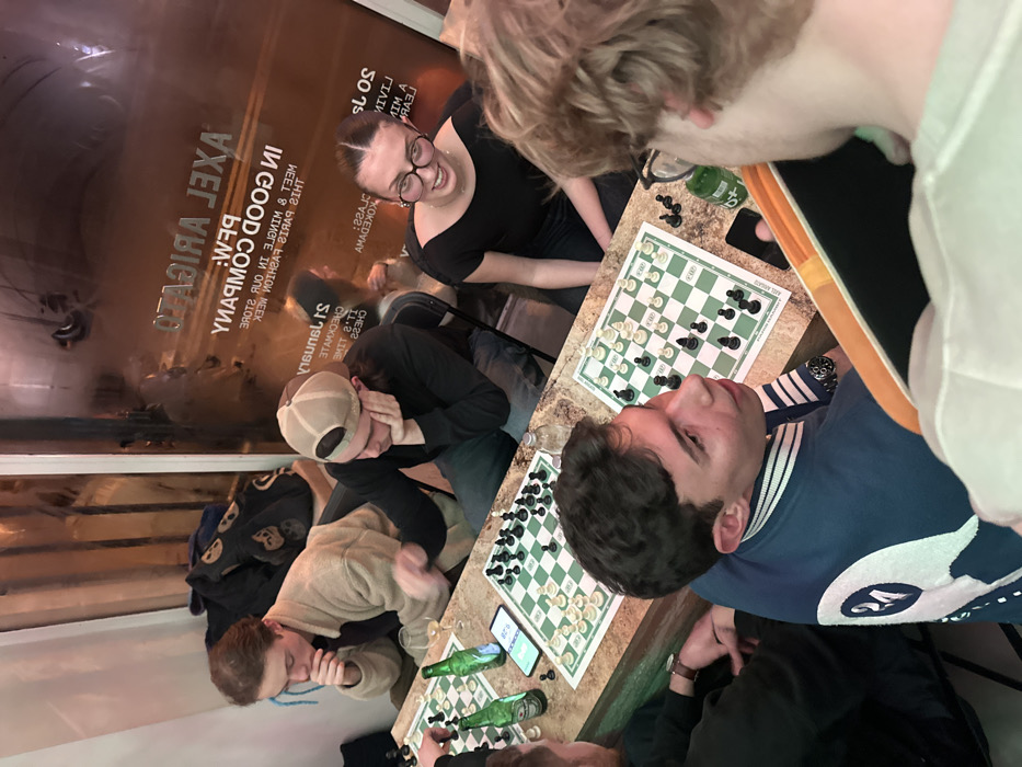 A group of people are playing chess. Three chessboards are visible on a table, each with a partly-played game in progress. Several green glass bottles are scattered around the boards. One person is using a phone or device next to their board. Text visible in the background reads "AXEL ARIGATO" and "IN GOOD COMPANY".