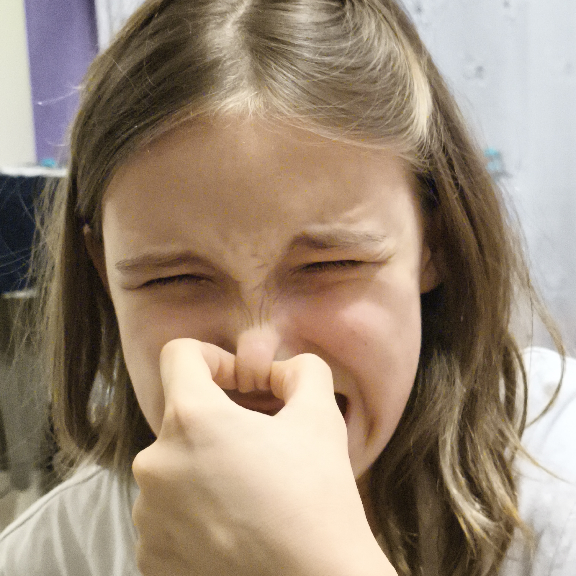 A young person with light brown hair holds their nose with their right hand. Their eyes are tightly closed, and their face is contorted as if reacting to a strong smell. The person wears a white shirt. The background is blurred, with hints of a wall, a cabinet, and possibly a piece of furniture covered with dark clothing. There is no definitive information to indicate the location in the city of Chaikovsky, Russia.