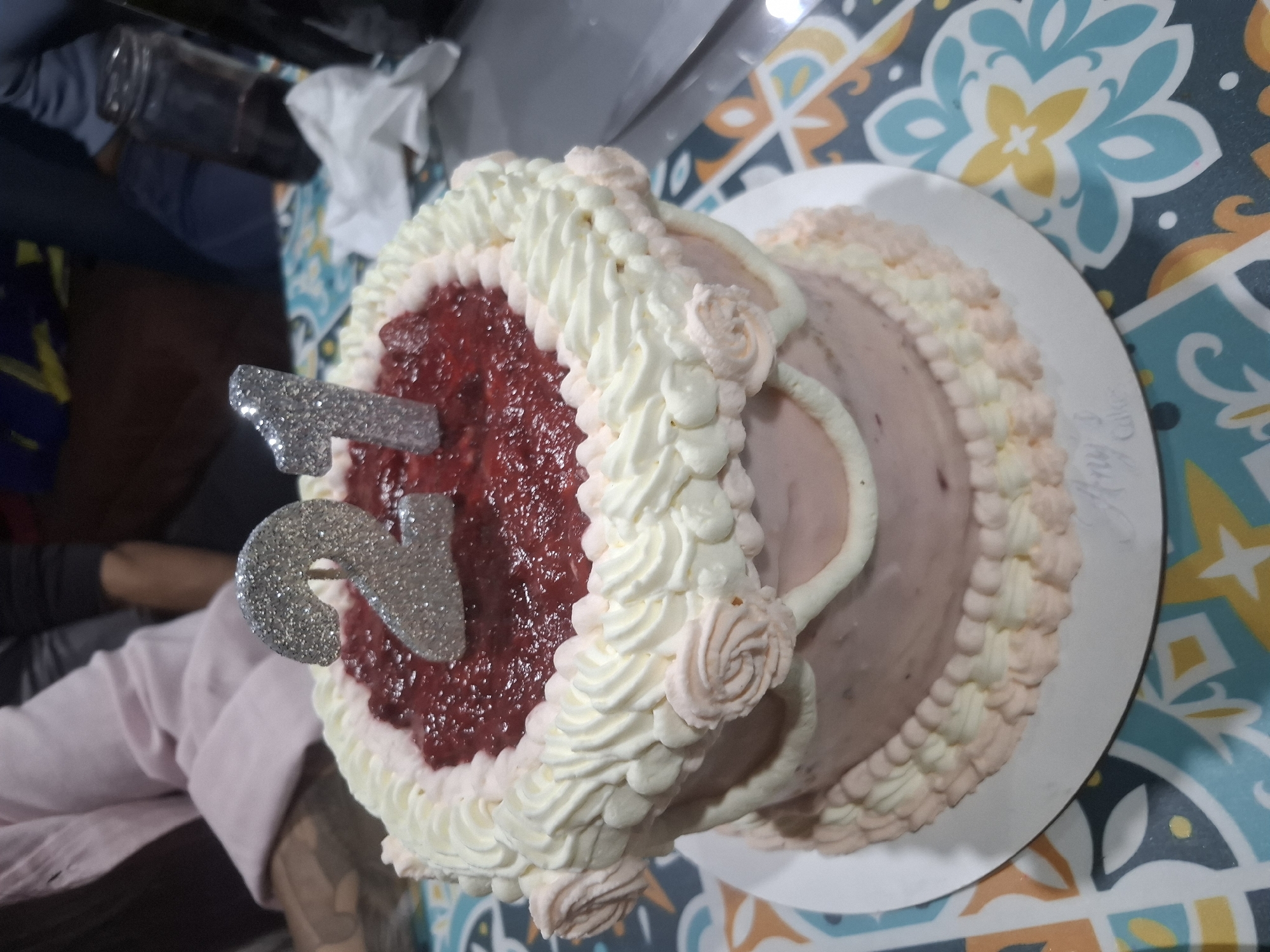 A close-up, overhead shot depicts a round cake on a white plate. The cake has a two-tone pink frosting with white and pink decorative swirls along the outer edges. One half of the cake is topped with a dark red, jam-like substance. Silver glittery number "21" cake toppers are inserted into the red topping. The cake is positioned on a table covered with a colorful patterned tablecloth. The lower left corner of the image shows a person's arm and torso, wearing a pink shirt. In the background, there's a glimpse of items, including what appears to be a glass jar and some white tissue. The location is likely a home or indoor setting, but the specific city of Buenos Aires, Argentina, cannot be determined from the visual data provided.