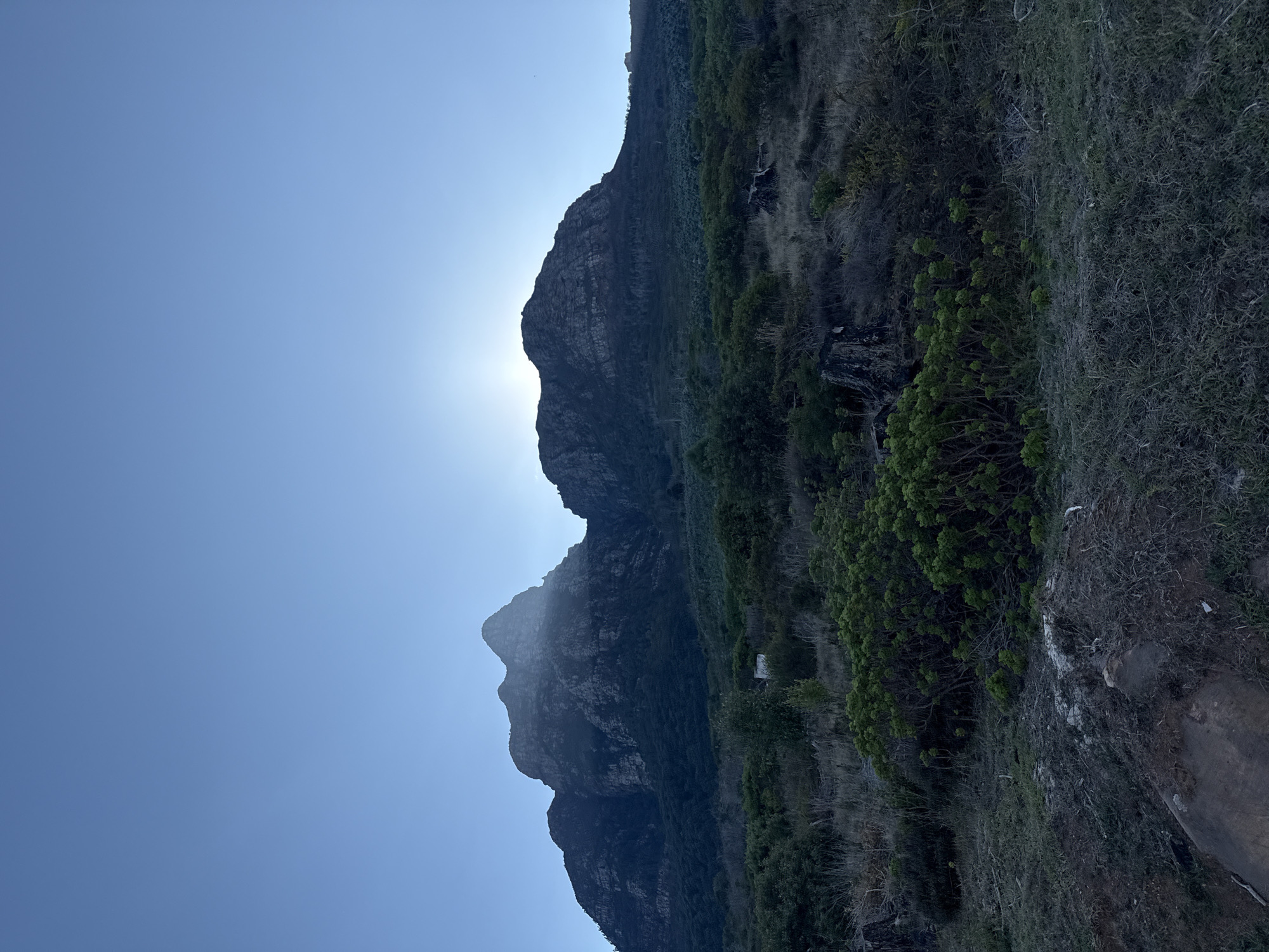 The image shows a mountainous landscape in Cape Town, South Africa, likely taken on a sunny day. Two distinct peaks are visible against a clear blue sky, with the sun positioned behind the higher peak, creating a backlight effect. The mountainside is covered in vegetation, including various shrubs and grassy areas. The foreground shows a rocky terrain.