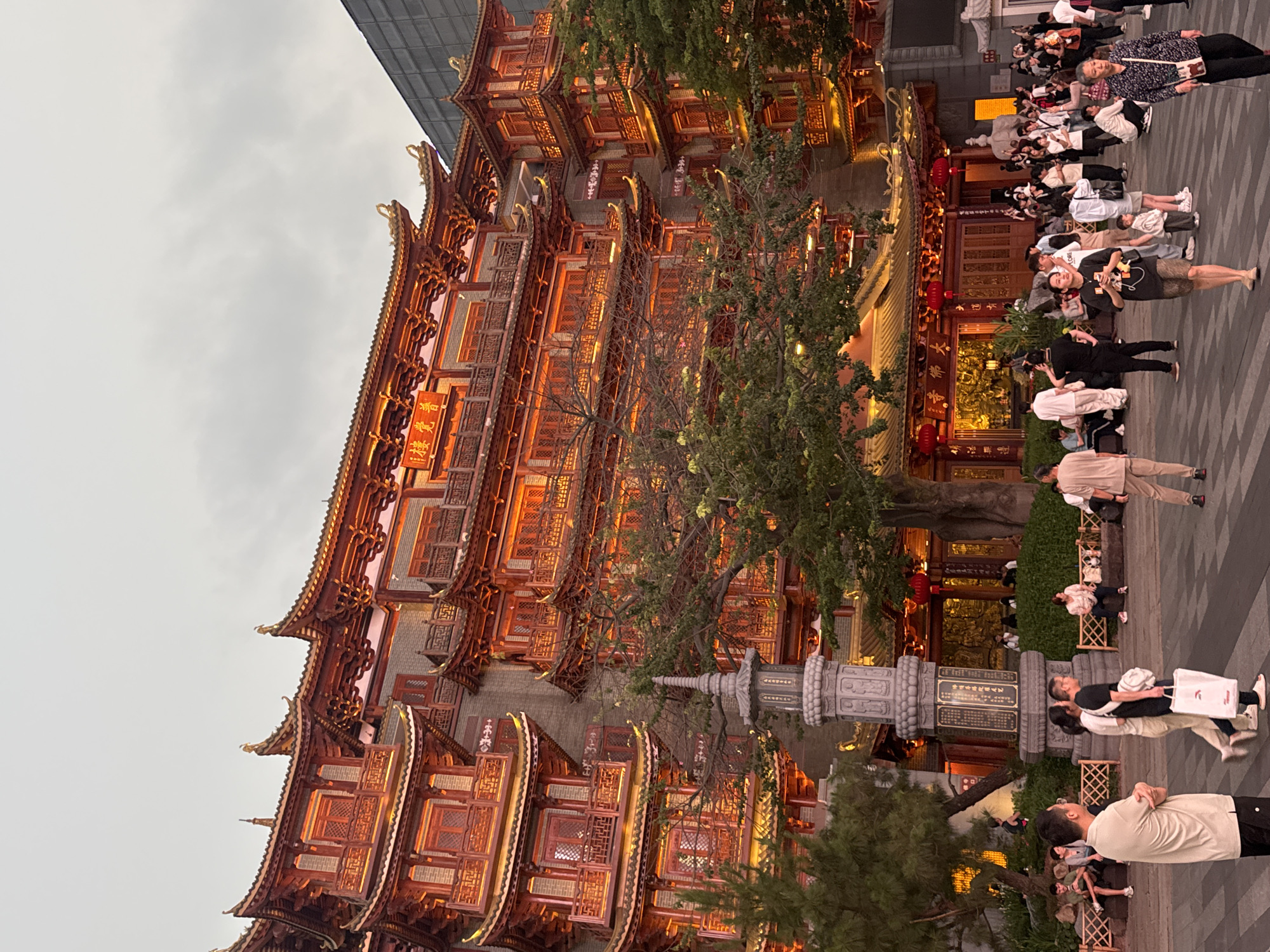 A vertical, medium shot depicts a building in Yuexiu District, China. The traditional red and brown wooden building has multiple layers of roofs and windows, illuminated with a warm orange glow. Trees partially obscure the facade. In the foreground, a stone monument stands on a paved area, where people are walking. The sky is overcast.