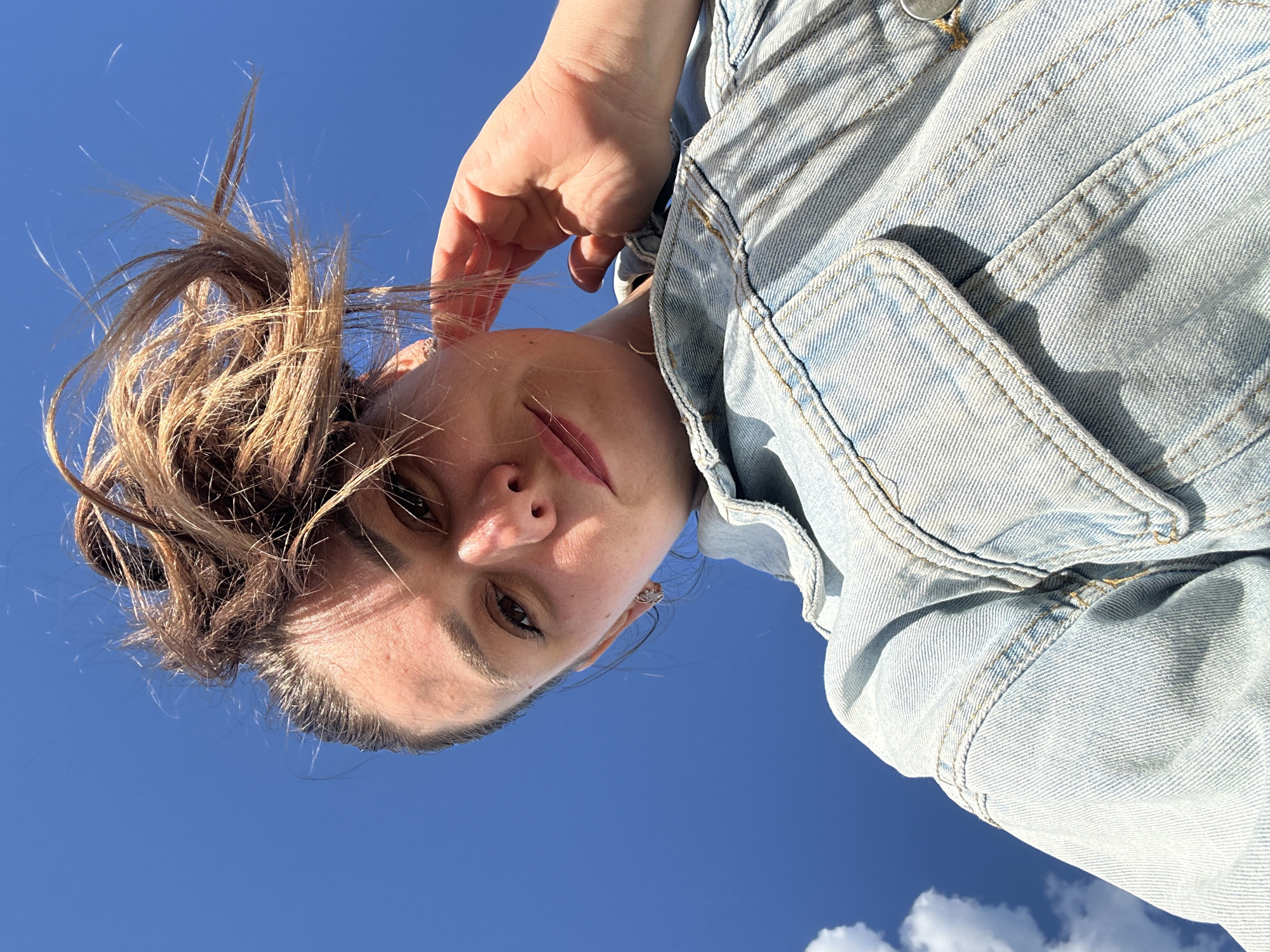 A woman is looking down at the camera. She is wearing a denim jacket. Her hair is pulled back in a messy bun. The sky is blue in the background. There are a few white clouds in the lower right corner. The location and city cannot be determined from the image.