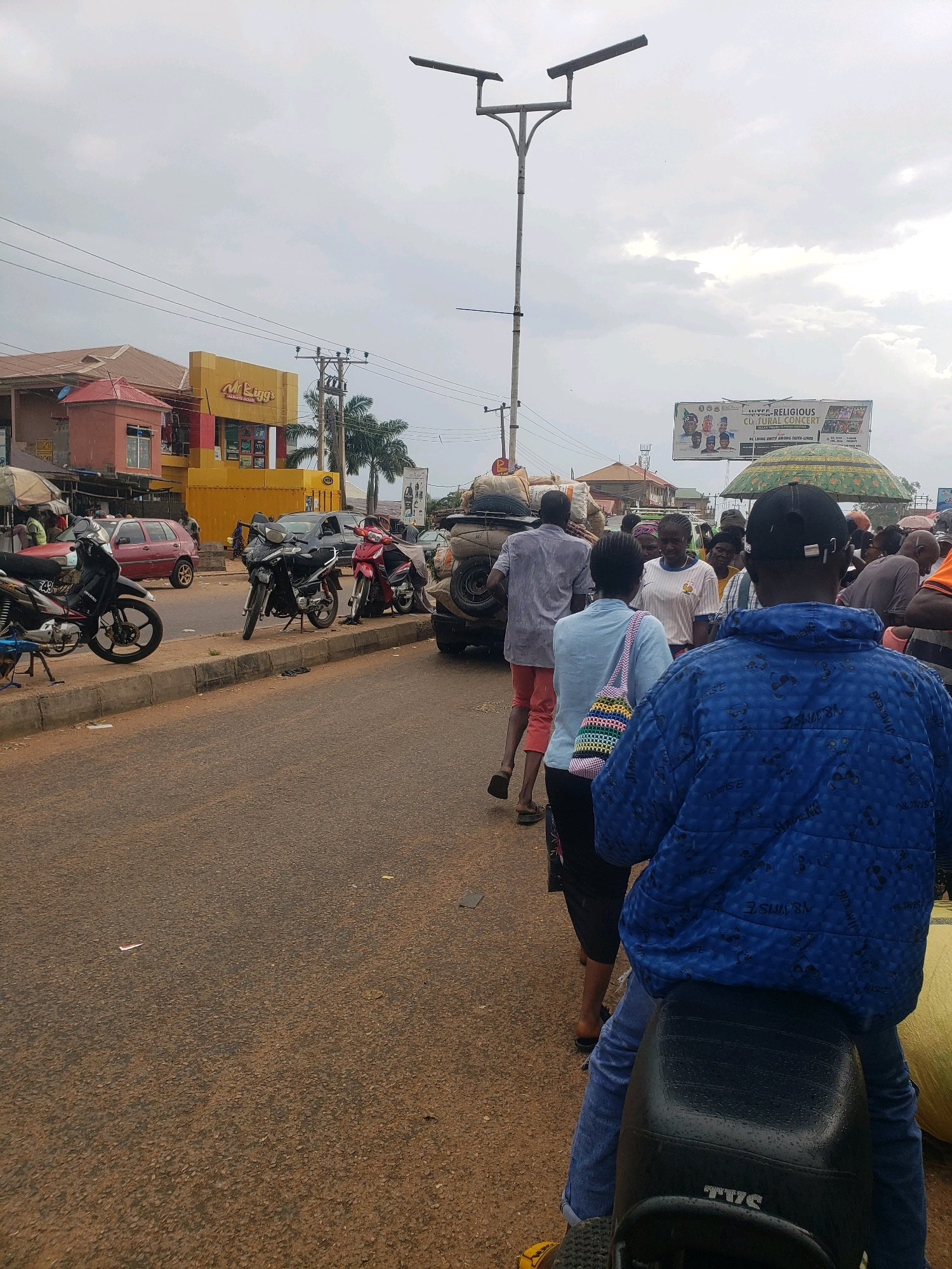 The image depicts a street scene in Sabon Tasha, Nigeria, under an overcast sky. In the foreground, a person wearing a blue patterned jacket and blue jeans is seated on a motorcycle, facing away from the camera. The motorcycle has a black seat with the brand name "TVS" visible. To the right of the motorcycle, a large, yellow object, possibly a bag of produce, is partially visible.
Behind the motorcycle, a group of people are walking along the side of the road. Several individuals are carrying bundles or bags on their heads or in their hands. One person in the middle distance, wearing a light blue shirt and carrying a colorful woven bag, is walking towards the right. Further back, a man in a grey shirt and red trousers is walking away from the camera.
Several motorcycles are parked along the curb. A red car is visible to the left, and a black car is parked further down the road. Buildings line the street, including a prominent yellow building with red trim, identified by signage as "Mr. Biggs." A billboard advertising a "united religious cultural concert" is visible in the background. A street lamp with two lights stands near the center of the frame. The road surface appears to be asphalt, with scattered debris. The overall impression is of a busy market or transport hub.