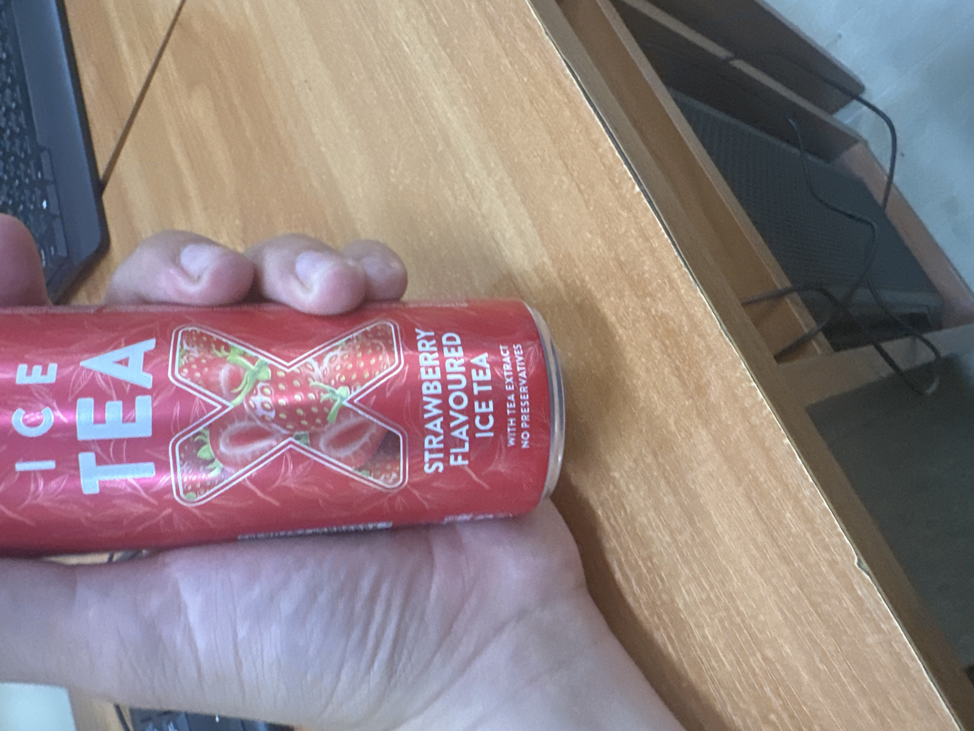 A person's hand holds a red aluminum can of "ICE TEA" strawberry-flavored drink on a wooden table. A keyboard is partially visible to the left of the hand. A wooden structure with black wiring is visible to the right. The scene is located inside, likely indoors near a desk. The location in Varna, Bulgaria, is not discernible from the image.