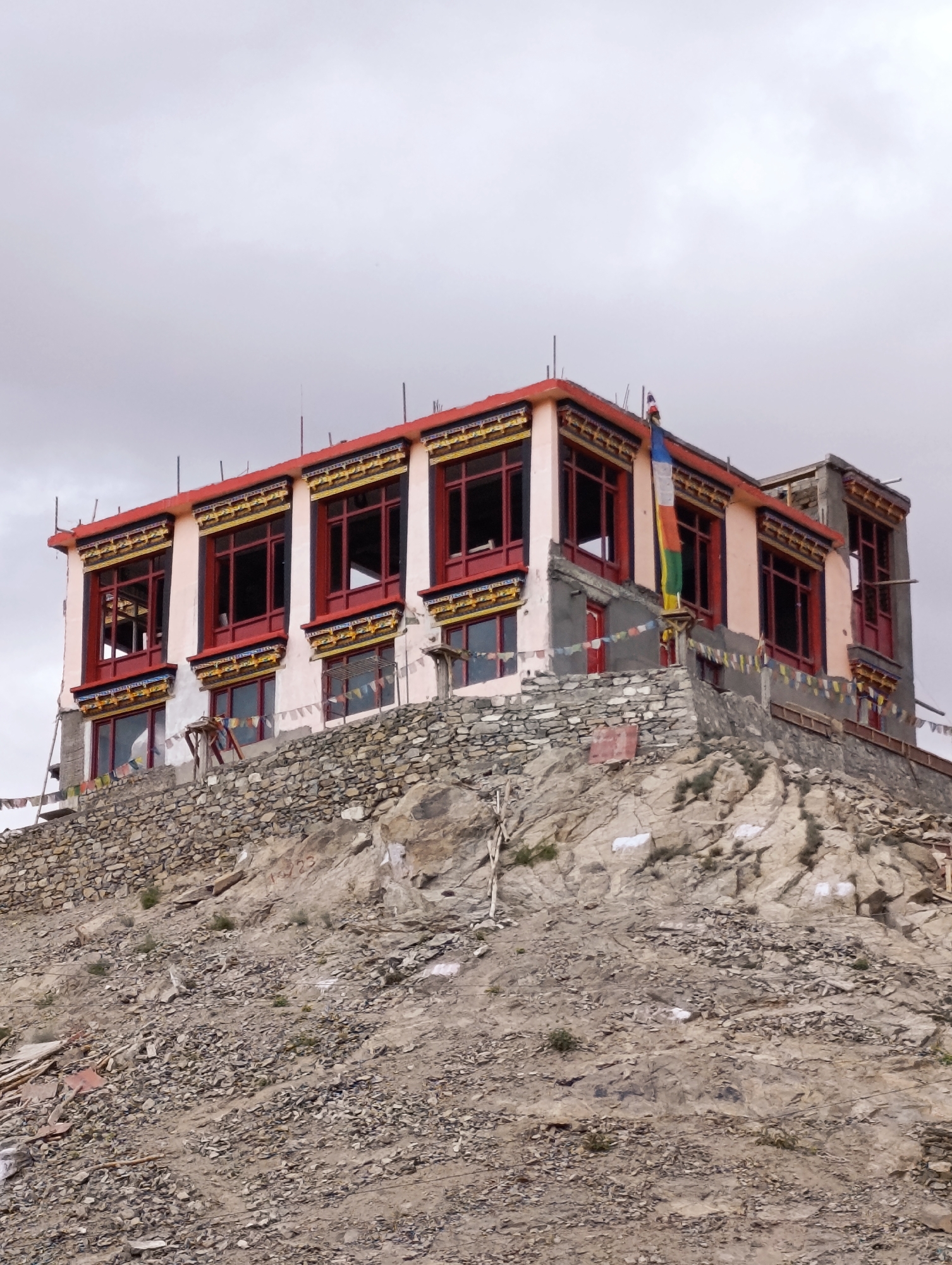 A multi-story building with a light pink facade and red-trimmed windows sits atop a rocky, debris-strewn hill under a cloudy sky. The building has multiple levels, each featuring rows of large windows with red frames. Decorative yellow and blue trim adorns the upper sections of the windows. Prayer flags in various colors are strung along the lower part of the building and on the hillside below. A Tibetan flag, composed of vertical stripes of blue, white, red, green, and yellow, hangs from a pole on the right side of the building. The structure appears to be undergoing construction or renovation, with some exposed concrete and scaffolding visible. The overall scene suggests a remote, mountainous location. The building is identifiable as the Sankar Gompa, a Buddhist monastery located in the city of Leh, India.