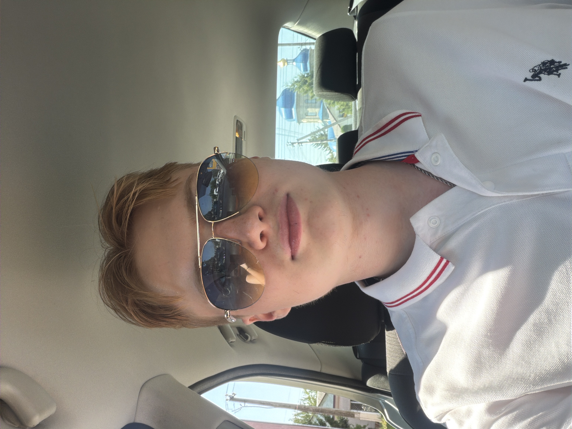 A young person with reddish-blond hair and sunglasses is taking a selfie inside a car. They are wearing a white polo shirt with red and blue trim on the collar. The car's interior is visible, including the headrest of the seat behind them. Through the car windows, a church with blue domes and a golden cross can be seen, suggesting the location is Polonne, Ukraine. The person's face is partially obscured by sunglasses. The lighting suggests it is daytime.