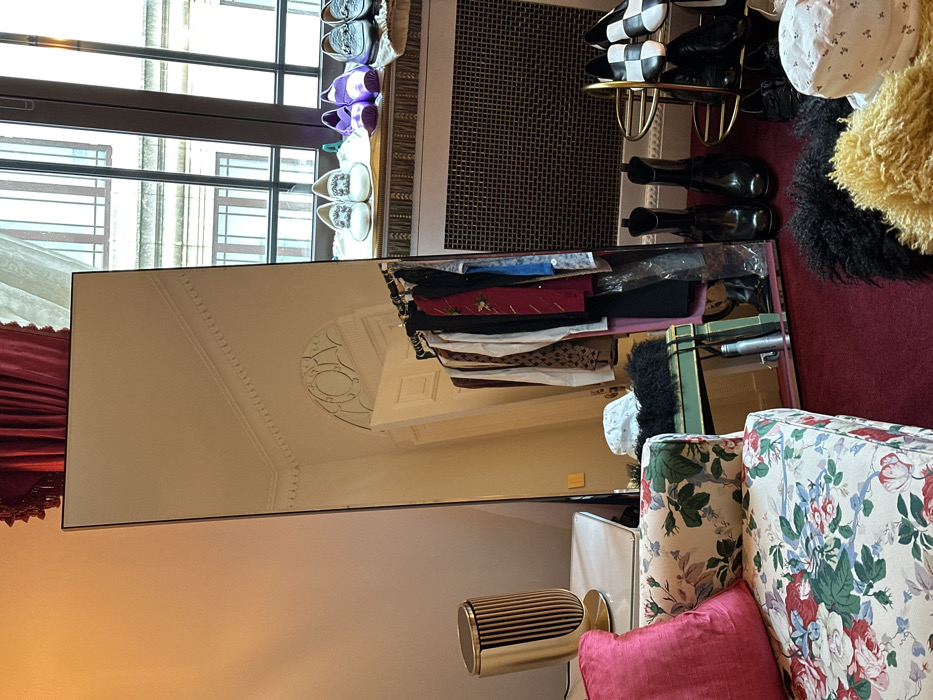 An indoor scene depicts a full-length mirror reflecting a room with classical architectural features, positioned within what appears to be a dressing area or bedroom in a building in London, United Kingdom.
The primary subject is a tall, rectangular, dark-framed mirror standing vertically, angled slightly right. Its reflection shows a white room with a high ceiling featuring decorative molding and an arched doorway with an ornate transom above. A clothing rack filled with various garments, including shirts, jackets, and dresses, is visible in the reflection to the right of the doorway. Below the clothing rack, a white patterned hat rests on a dark, textured object, which sits on a green surface. A silver water bottle is reflected on the floor near the mirror's base.
To the left of the mirror, a portion of a floral-patterned sofa or armchair with a solid pink cushion is visible in the foreground. Adjacent to the sofa is a white table or stand supporting a gold, ribbed, cylindrical object, possibly a speaker or decorative lamp. The wall in this area is a light, warm tone. Red velvet curtains with tassels are visible in the upper left corner, partially obscuring the top of the mirror.
To the right of the mirror, a large window with multiple vertical panes and dark frames overlooks a light-colored, multi-story building with similar window structures, indicative of urban architecture. Below the window is a metal radiator with a gridded cover. On the windowsill, three pairs of shoes are arranged: white embellished loafers, purple ballet flats, and dark embellished loafers.
On the red carpeted floor in the foreground, two pairs of black, glossy, tall boots stand near the base of the mirror. A gold-toned, multi-tiered shoe rack displays several pairs of shoes, including black and white checkered flats and black heels. Two textured, fur-like objects—one black, one yellow/light brown—are present on the floor, alongside a white patterned bucket hat. No individuals are present in the scene. The architectural details, both interior and exterior, strongly suggest a location within an older, substantial building in London.
