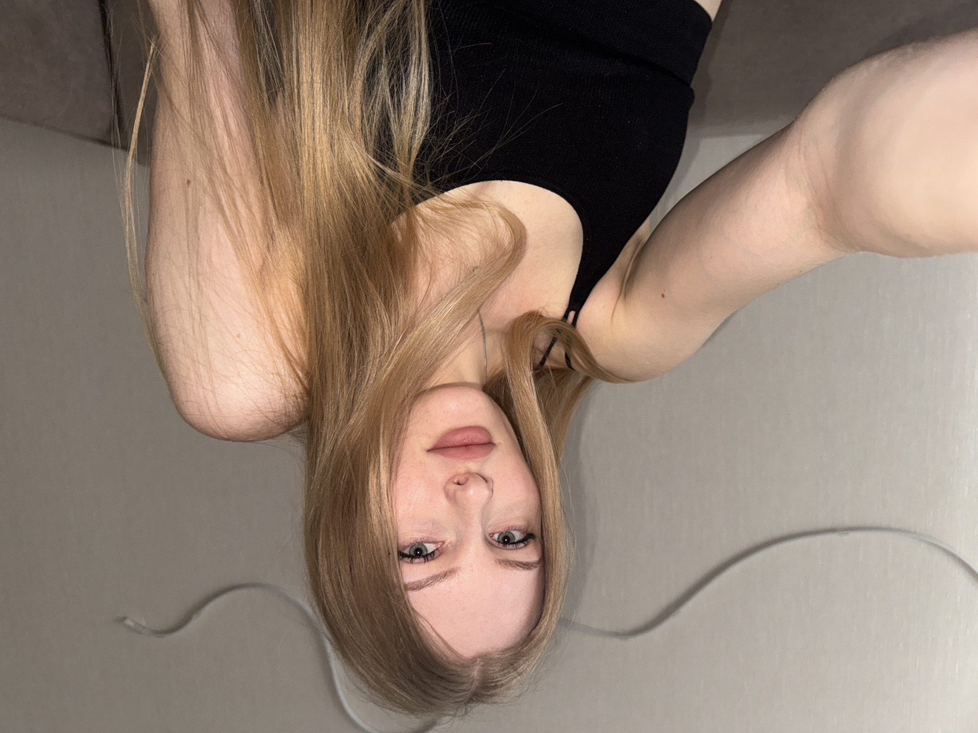 A selfie of a woman is captured indoors. The woman has long, blonde hair and is wearing a black top. She is situated in what appears to be a room with a plain wall in the background. The image is presented in an upside-down orientation. The location is not explicitly verifiable as Perm, Russia, from the image alone.