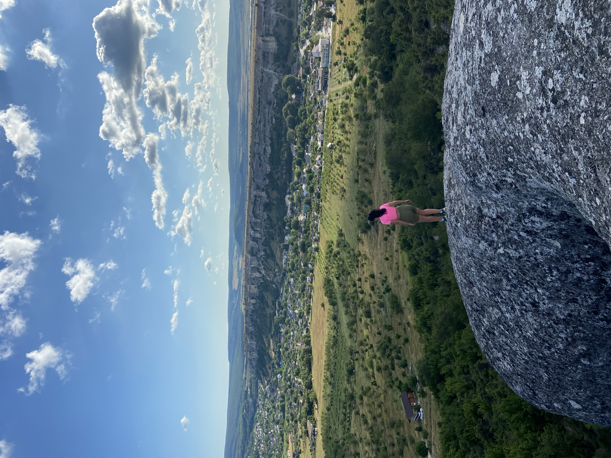 In the city of Krasnomakskoye, Ukraine, a woman stands on a large rock overlooking a valley. She is wearing a pink crop top and green shorts. Below her, a village is nestled among rolling green hills. In the background, a long, flat-topped mountain rises, its rocky face illuminated by the sun. The sky above is a clear blue, dotted with fluffy white clouds. The scene is one of natural beauty and tranquility, with the woman appearing to be enjoying the panoramic view.