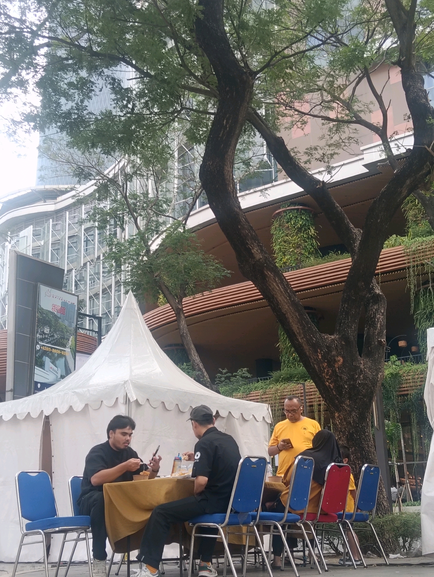 In South Jakarta, Indonesia, a group of people are seated at tables set up outside a white tent, with a large tree and a modern building in the background. A man in a black shirt is eating and looking at his phone. Another man in a black shirt and cap is also eating. A man in a yellow shirt is standing and looking at his phone, and a woman in a black headscarf is seated. The environment suggests an outdoor dining or event setup, with blue and red chairs around tables covered in tan tablecloths. The building in the background features glass facades and tiered balconies with greenery.