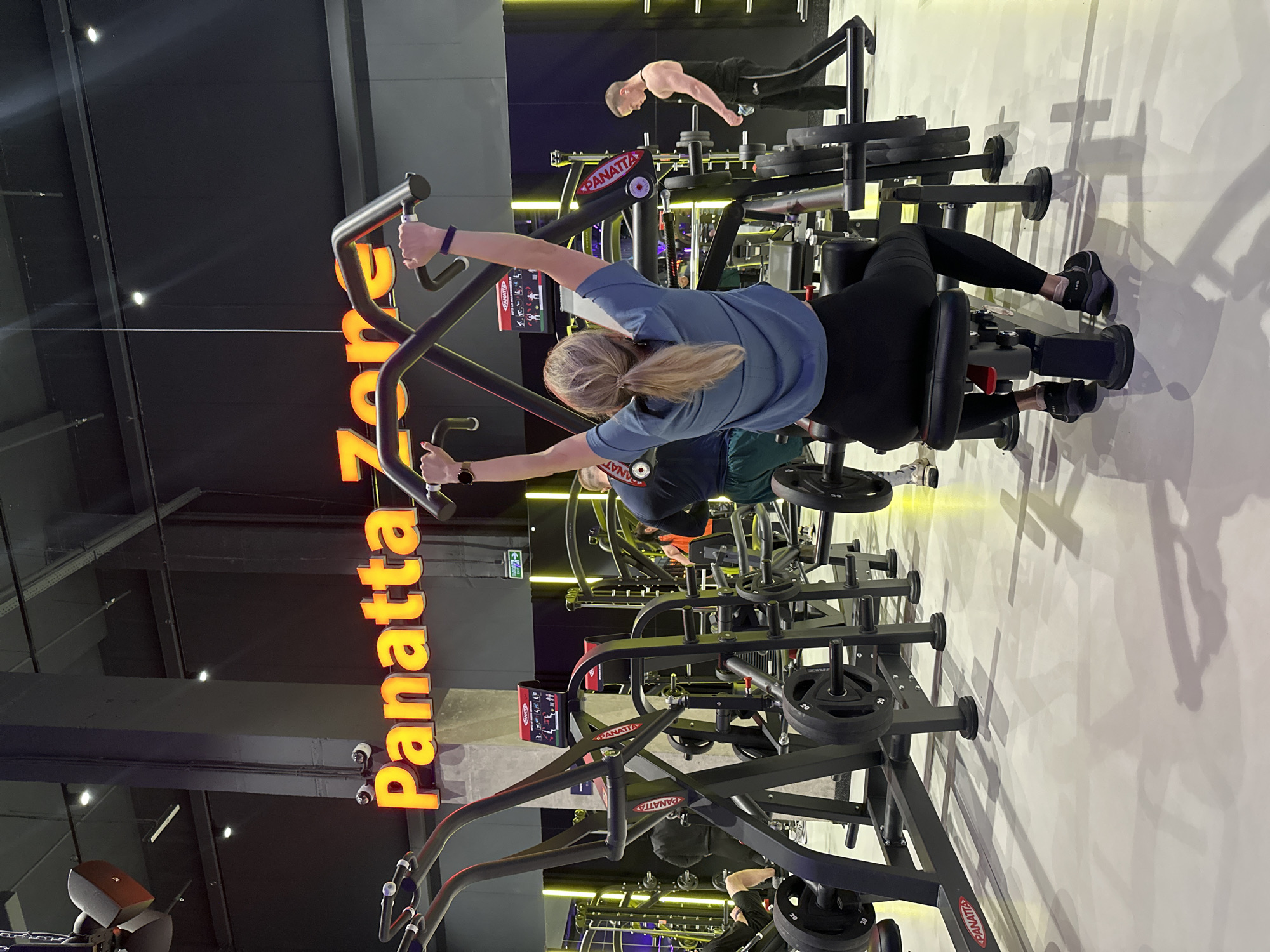 The image captures the interior of a modern gym, identified as Panatta Zone in Murino, Russia. In the foreground, a woman with blonde hair pulled back in a ponytail is seated on a weight machine, performing an exercise with her arms raised and holding onto the machine's handles. She is wearing a blue t-shirt and black leggings. Behind her, other gym-goers are visible, including a man in the background to the right wearing a black tank top and black pants, and a person in the mid-ground to the left wearing a dark shirt and green shorts. Various pieces of gym equipment, including weight machines and barbells, are arranged throughout the space. The gym features a brightly lit environment with a prominent neon sign spelling "Panatta Zone" in orange lettering on the wall. The flooring is light-colored and reflects the lighting, casting shadows from the equipment. The overall scene depicts a typical gym environment with individuals engaged in physical activity.