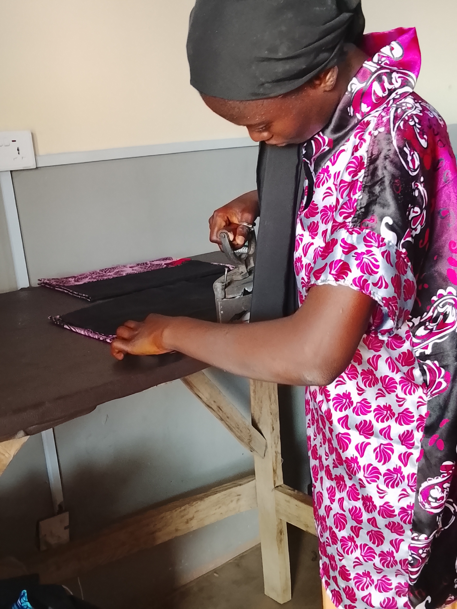 A person is shown in a close-up, indoor shot, intently ironing fabric. The individual, likely female, has dark skin and is wearing a black head covering and a brightly patterned garment featuring a mix of pink, white, and black floral or abstract designs. She is leaning over a rustic, dark brown wooden table, which serves as an ironing surface.
In her right hand, she holds a heavy, dark-colored, old-fashioned iron with a handle, pressing it onto a piece of black fabric. Her left hand is placed on the fabric, guiding it. Another folded piece of fabric, matching the pattern of her garment, is visible on the table to the left. The person's hands show a reddish-brown discoloration, possibly from henna or a working substance.
The background consists of a light-colored (beige or cream) wall above a gray dado rail. A white electrical outlet with two sockets is mounted on the wall to the left. The wooden table is supported by visible wooden beams. The overall scene suggests a functional workspace, possibly in a home or small workshop, consistent with daily activities in Akwanga, Nigeria. The lighting appears natural, and the person is focused on her task. There is no visible text in the image.