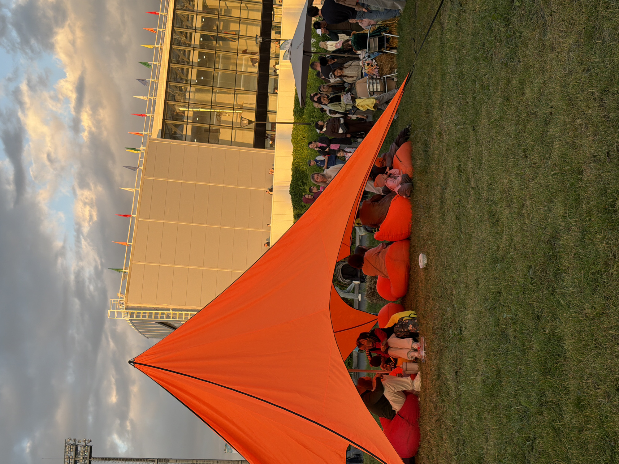 The image depicts an outdoor gathering on a grassy field, likely during the late afternoon or early evening, given the warm, golden light illuminating the scene and the dramatic clouds in the sky. In the foreground, a large, bright orange tent or canopy is pitched, providing shade for a group of people seated on large, colorful beanbag chairs.
Beneath the orange canopy, several individuals are visible. Two people are seated close together on red beanbags, one wearing a dark jacket and white pants, the other a dark top and light-colored pants, appearing to be in conversation. To their right, another person is seated on a red beanbag, facing away from the camera. More beanbag chairs are scattered around, some occupied by other individuals, including a child in a pink outfit. A small white bowl is on the ground near the beanbags.
Behind the canopy, a larger group of people are standing and mingling. Some are gathered around hay bales, suggesting a rustic or festival-like atmosphere. There's a large, grey umbrella providing shade for some of these individuals. The background features a modern, large building with a glass facade and light-colored exterior walls. Flags are visible along the top edge of this building. To the left of the building, a tall stadium light structure is present. The overall environment is a park or open space adjacent to a modern structure, with a clear sky with prominent cloud formations. The grass is green and well-maintained.