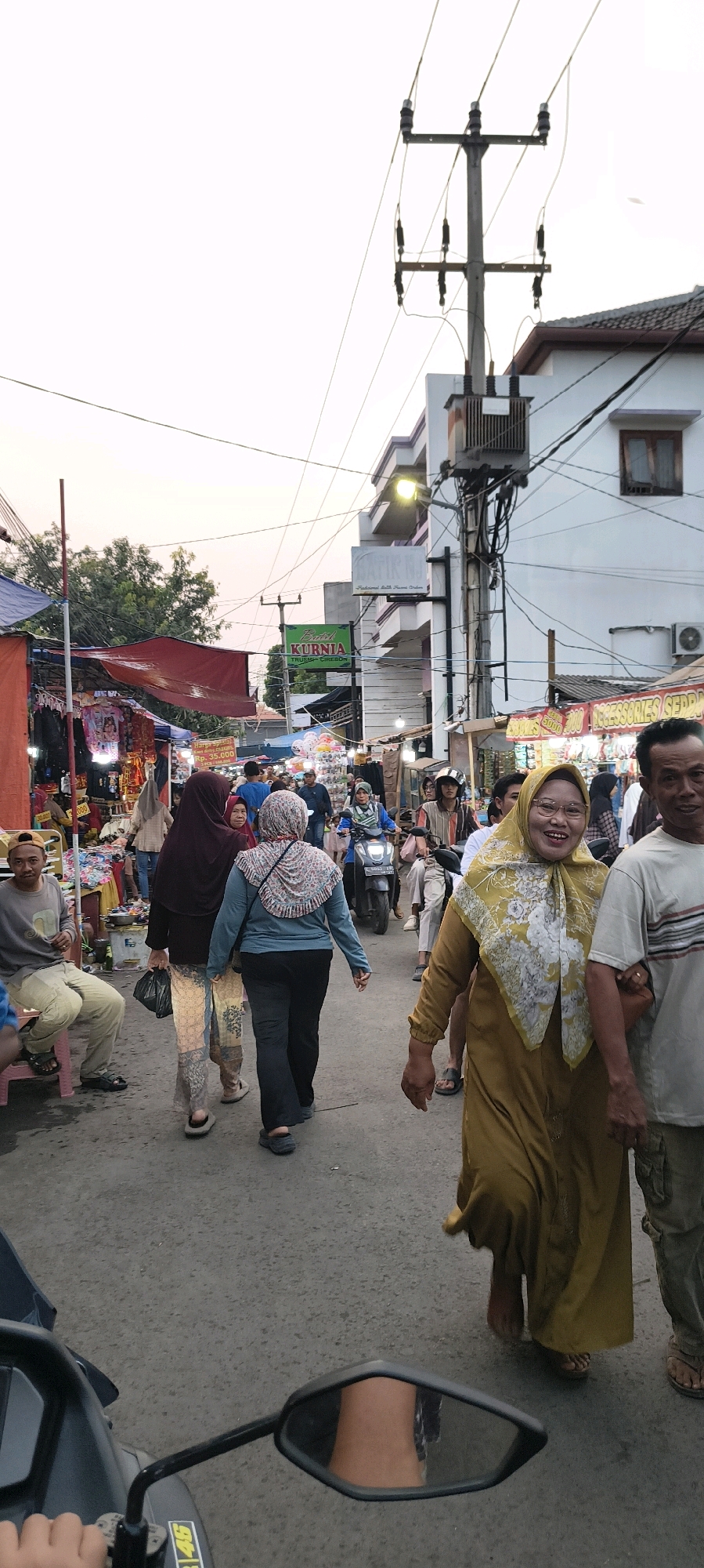 This is a candid, eye-level, vertical shot captured from the perspective of a person on a motorcycle in Trusmi Wetan, Indonesia. The scene depicts a bustling street market, possibly during late afternoon or early evening, as indicated by the soft lighting and the overhead power lines. In the foreground, a motorcycle's handlebar and rearview mirror are visible. The mirror reflects a portion of the rider's leg and the street behind them.
The street is moderately crowded with people. Dominating the right side of the frame is a smiling woman wearing a yellow hijab and a matching long dress, adorned with a floral pattern. She is walking towards the viewer, with her right arm slightly bent and her hand resting near her hip. To her right, an older man in a light-colored, striped t-shirt and khaki pants is walking.
In the mid-ground, a woman in a blue hijab and a light blue top with a patterned scarf walks away from the camera, followed by another woman in a dark maroon hijab and a patterned skirt. Other individuals, some on foot and one on a motorcycle, are also present amidst the market stalls.
The market stalls are set up on both sides of the street, with awnings and tarps providing shade. Various goods, including clothing and accessories, are displayed. Signs are visible, including one for "BATIK KURNIA" with "TRUSMI - CIREBON" below it, confirming the location. Electrical poles and transformers are prominent features of the urban environment. The overall atmosphere suggests a lively local marketplace.