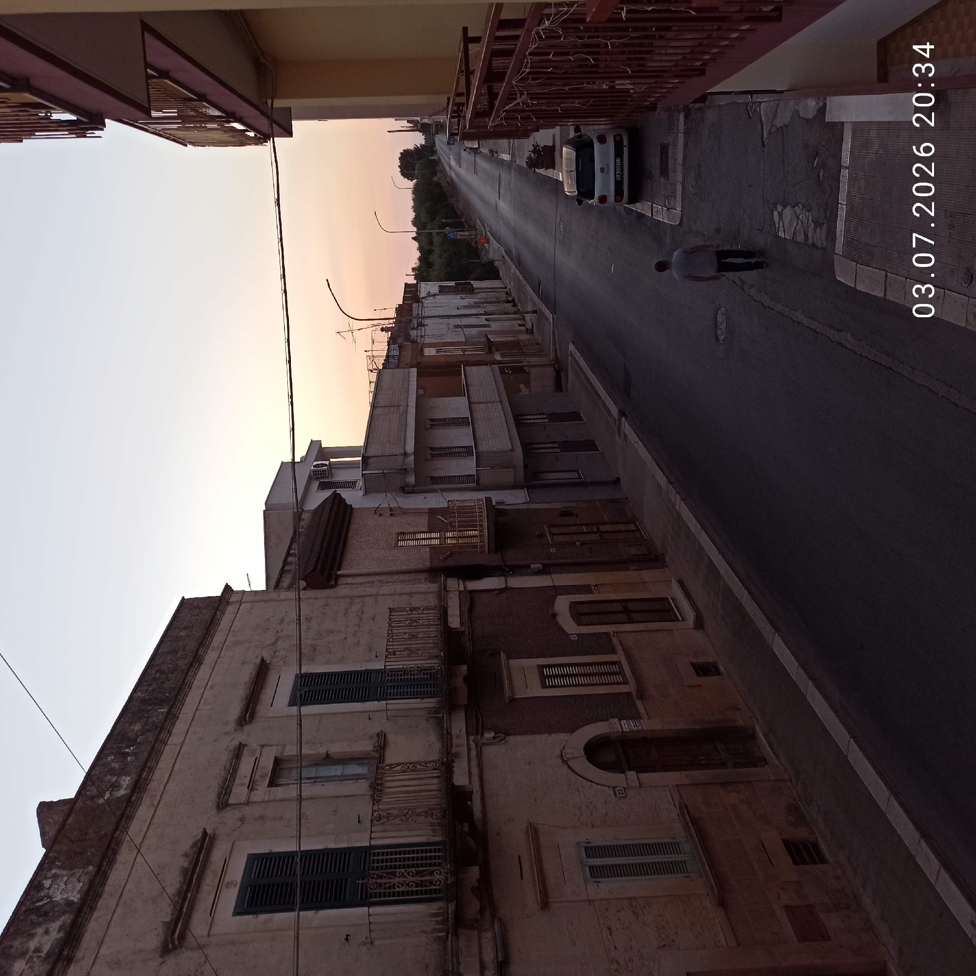 The image depicts a street scene at dusk in Bitonto, Italy. A man is walking away from the camera down a paved street lined with multi-story buildings. The buildings on the left appear older, with weathered facades and wrought-iron balconies. The buildings on the right are more modern, with balconies and yellow and red colored exteriors. A white compact car is parked on the right side of the street. The sky is a gradient of pale orange and blue, indicating the sun has recently set. The timestamp "03.07.2026 20:34" is visible in the bottom right corner of the image. The scene appears to be residential, with street lights visible in the distance.