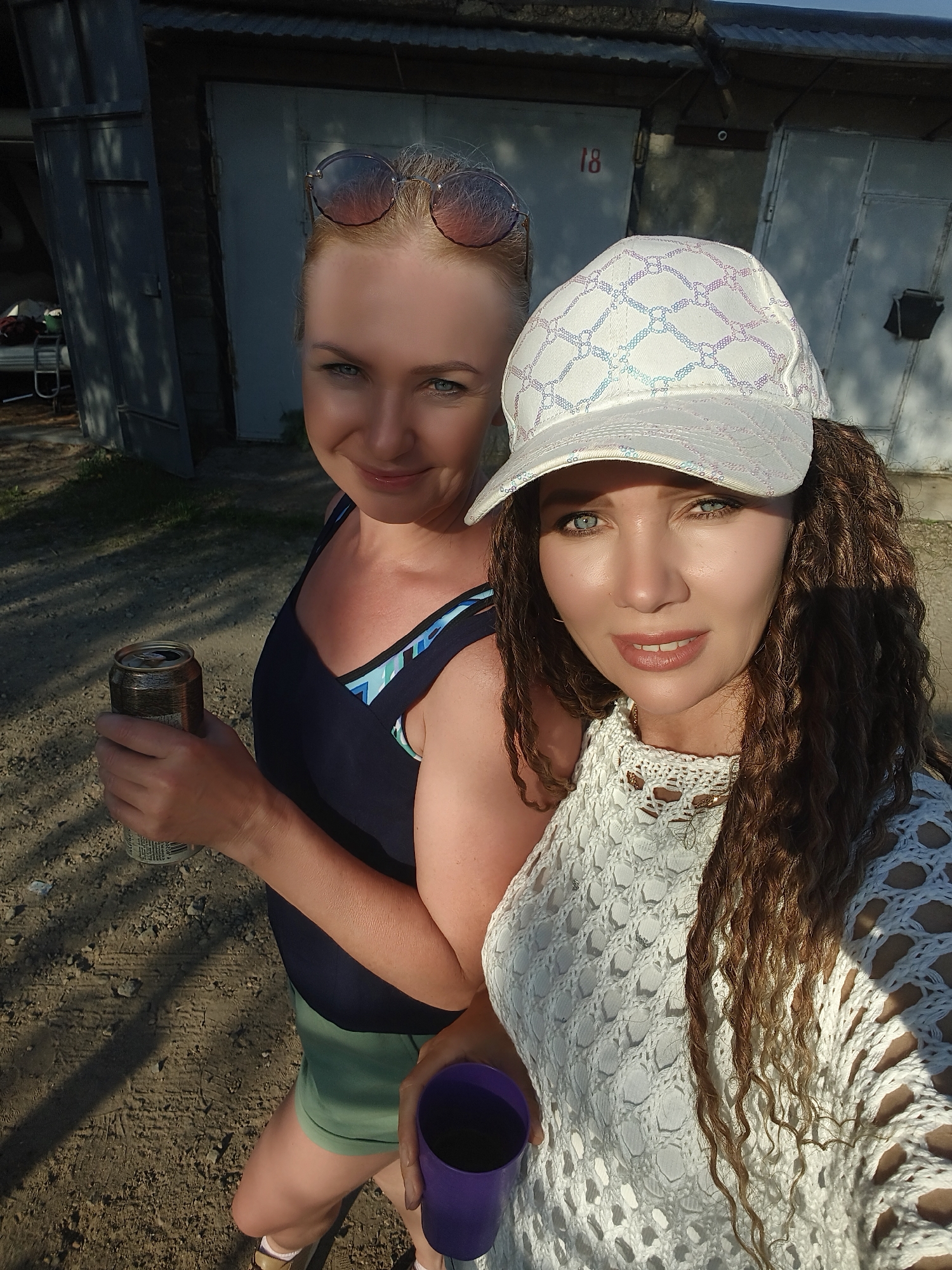 The image shows two women standing outside in front of a row of garage doors. The garage door closest to the camera is labeled "18". The woman on the left has blonde hair pulled back, sunglasses on her head, and is wearing a dark blue tank top and green shorts. She is holding a beer can in her left hand. The woman on the right has curly brown hair and is wearing a white patterned baseball cap and a white crocheted top. She is holding a purple cup in her left hand. The ground is dirt and gravel. The location appears to be in the city of Unknown, Unknown.