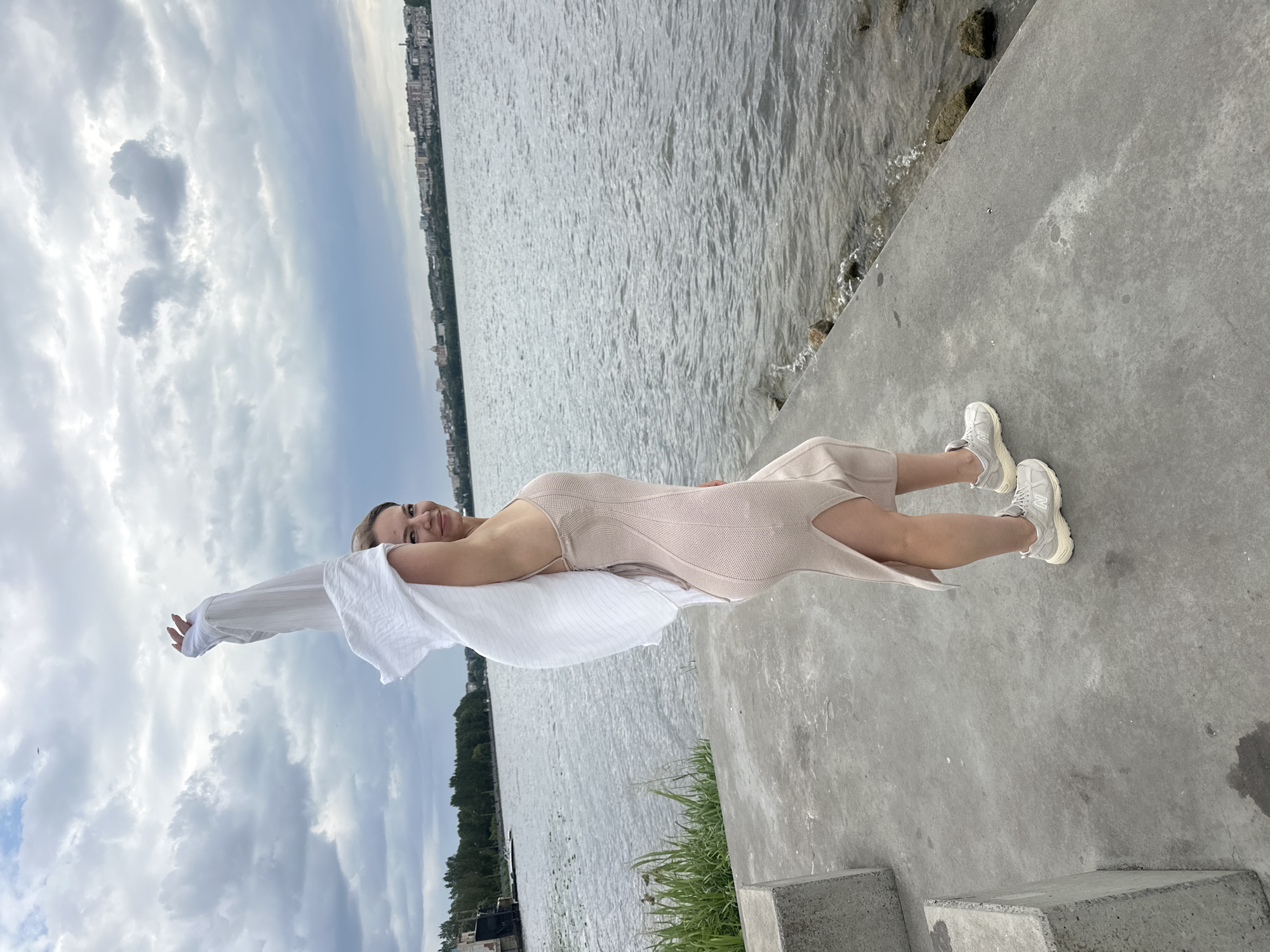 A woman stands on a concrete platform near a body of water, holding a white shirt over her head. She is wearing a light-colored, form-fitting dress with a thigh-high slit and white athletic shoes. Behind her, the water stretches out to meet a city skyline in the distance under a cloudy sky. The concrete platform appears to be part of a waterfront promenade or park, with some angular concrete structures visible to her left. The scene is likely Ternopil, Ukraine, given the presence of the Ternopil Pond, a large reservoir in the city center.