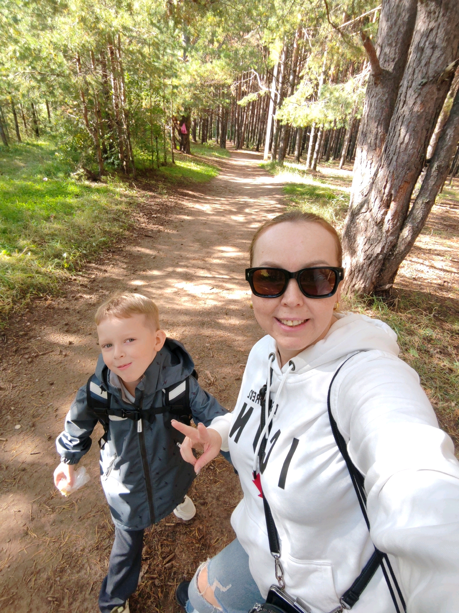 A woman and a young boy are taking a selfie in a park in Chaikovsky, Russia. The woman is wearing a white hoodie and sunglasses, while the boy is wearing a grey jacket and backpack. They are standing on a dirt path lined with trees. The sun is shining through the trees, casting shadows on the ground. The boy is holding a piece of food wrapped in plastic. The woman is making a peace sign with her fingers.