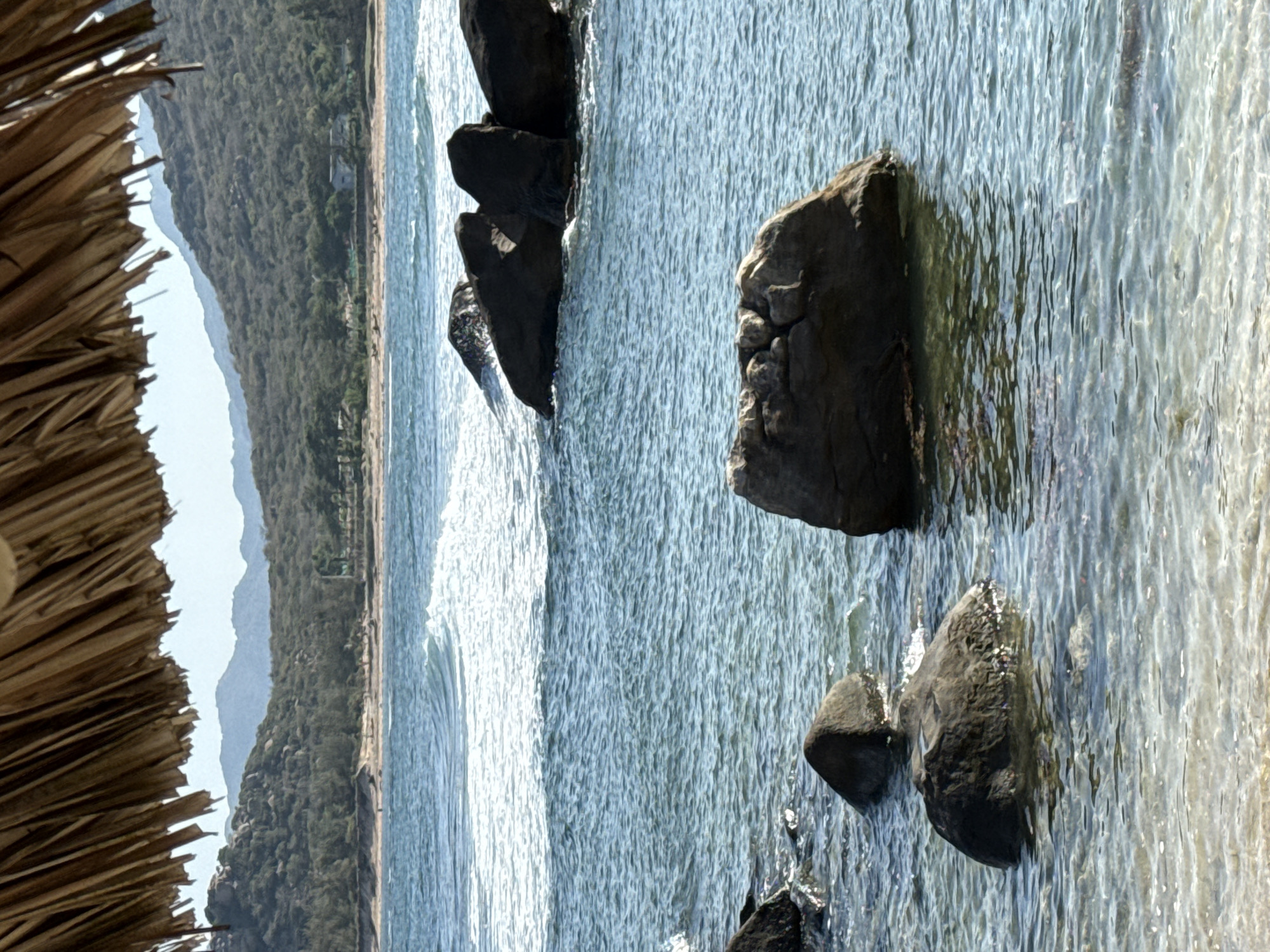 The image shows a view of the ocean with rocks in the water, possibly in Vinh Hai, Vietnam, based on the prompt. A thatch umbrella structure is visible in the left foreground. In the background, there is a coastline with lush, green vegetation and hills. The water is choppy and appears to be shallow near the shore. There are no people visible in the image. The scene suggests a coastal area with a rocky beach and clear, shallow water.