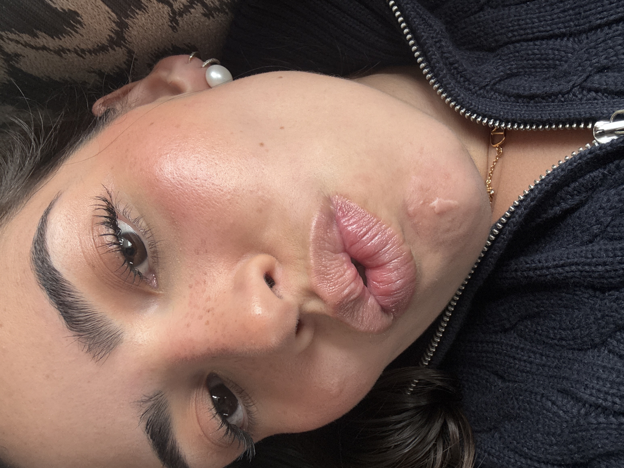 A close-up, slightly angled portrait features a person with fair skin, prominent cheekbones, full lips, and dark hair. The person's face is turned slightly towards the camera, with lips pursed in a kissing expression. The person has dark eyebrows and long eyelashes, and their eyes are a dark brown color. Some freckles are visible on their nose and cheeks. They are wearing a ring with a large pearl on their finger. The person is wearing a dark-colored zip-up knitted sweater, a gold chain is visible under the zipper. On the right cheek, near the jawline, there is what appears to be skin irritation or a small scar.
There is no information to suggest this image was taken in San Bernardino Tlaxcalancingo, Mexico.