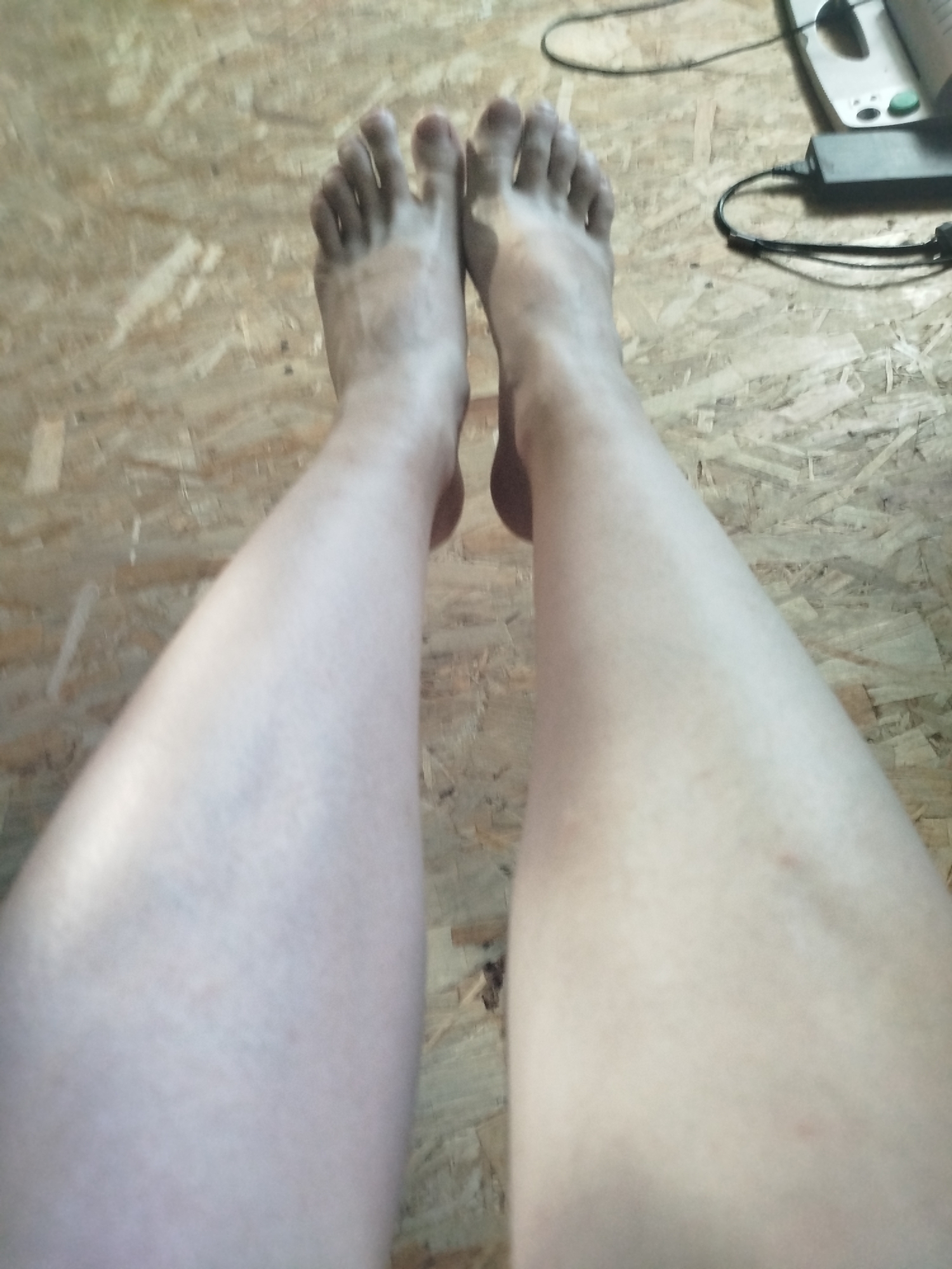 The image shows a first-person perspective of a person's bare legs and feet, positioned on a wooden floor. The floor appears to be made of particleboard or plywood. There are some electronic devices visible in the upper right corner of the image, including a black power adapter and what appears to be a book or manual. The lighting suggests an indoor environment. There are no discernible landmarks or clues to indicate the location in Zavodoukovsk, Russia.