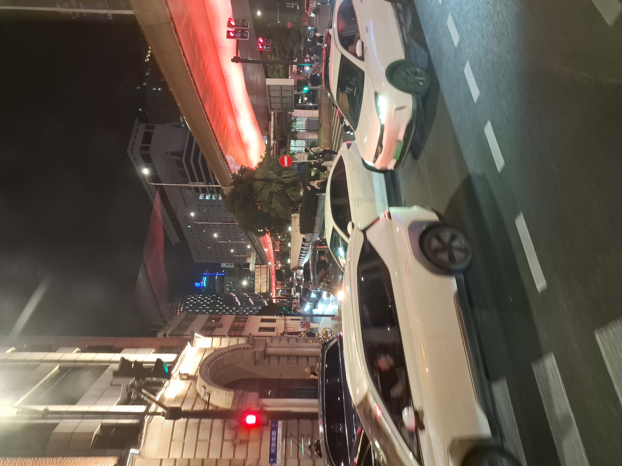 The image depicts a busy nighttime street scene in Shanghai, China. The foreground is dominated by several white SUVs, one of which is in sharp focus, showing its front end and the driver's side. The vehicles are on a road with white lane markings and a pedestrian crossing. In the background, there are buildings with illuminated windows, a red-lit overpass or bridge structure, and traffic lights. One traffic light shows red, indicating a stop. Several individuals are visible on the sidewalk and crossing the street. The overall impression is of urban traffic congestion at night.