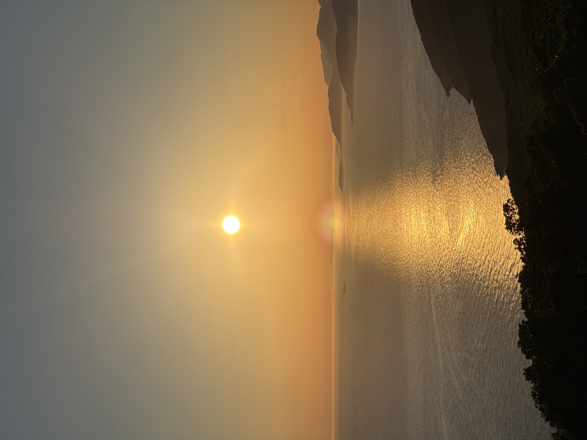The image depicts a serene sunset over the sea in Kovačko Polje, Montenegro. The sun, a bright orb, hangs low in the sky, casting a warm golden glow that reflects vibrantly on the water's surface. A faint trail in the sky suggests the passage of an aircraft. In the distance, a large ship is visible on the horizon. The coastline is characterized by undulating hills and mountains, silhouetted against the fading light. In the foreground, a dark, dense line of trees lines the edge of the land, adding a sense of depth to the composition. The overall atmosphere is peaceful and tranquil.
