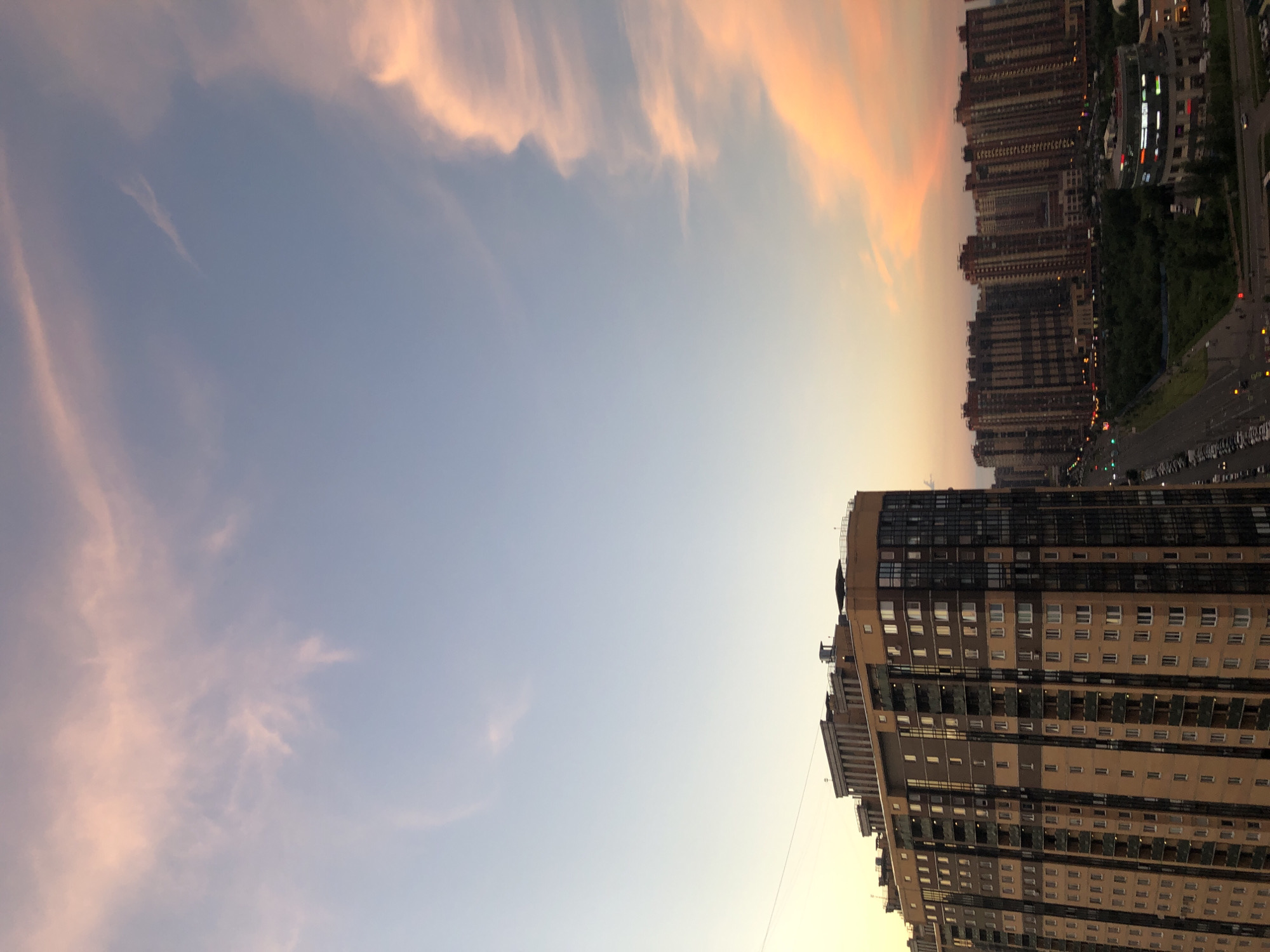 This is a vertical, eye-level, outdoor photograph of a city skyline at dusk. The sky is a pale blue with streaks of pink and orange clouds, illuminated by the setting sun. In the foreground, a tall, multi-story residential building with a tan and brown facade dominates the left side of the frame. The building features numerous windows and balconies. Beyond this building, a series of other apartment complexes stretch across the horizon. A road with cars and streetlights is visible in the midground, bordered by a strip of green trees and a circular, modern building with illuminated signage. The overall scene depicts a modern urban environment during the golden hour.