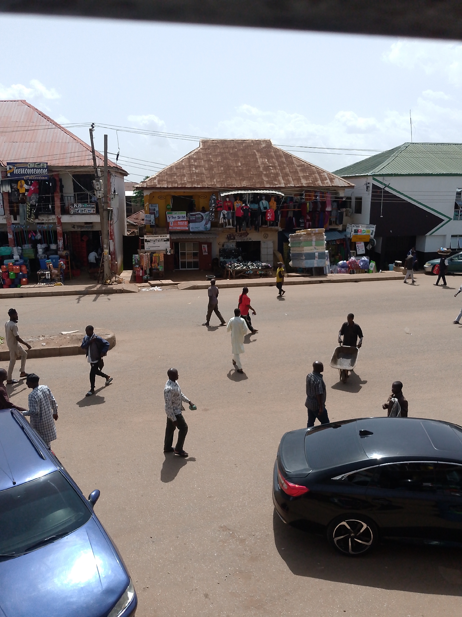This is an eye-level, medium shot of a bustling street scene in Bukuru, Nigeria. The image captures a busy intersection with several people walking and conducting business. The foreground is dominated by the hoods of two cars, a blue one on the left and a black one on the right. The street is paved with asphalt and shows signs of wear.
On the left side of the street, a two-story building with a red, corrugated metal roof houses several shops. The lower level displays a variety of goods, including what appear to be clothing items on mannequins and stacked merchandise. A sign above one shop reads "Phenomenon Collection."
Across the street, another building with a rusty, corrugated metal roof also houses shops. These shops seem to be selling various items, including what looks like mattresses stacked outside. Further down the street, a modern building with a green roof and white walls is visible. Signs indicating "Young Communications" and "FREE SIM" are displayed on one of the shops.
Several individuals are walking on the street. One man is pushing a wheelbarrow. Another man is carrying a large object on his head. Others are seen walking individually or in small groups, dressed in casual attire. The overall atmosphere suggests a busy marketplace or commercial area. The sky is partly cloudy, indicating daytime.