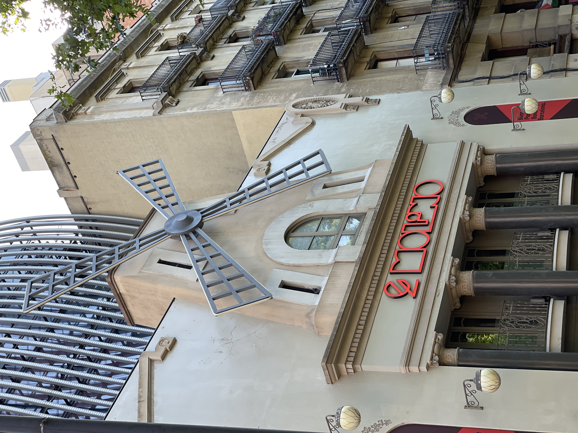 The image captures a worm's-eye view of a multi-story building facade in Barcelona, Spain, under bright daylight. The light-colored (beige/cream) building, viewed from below looking upwards, exhibits a blend of architectural styles.
A prominent feature on the lower part of the facade is a large, decorative metal structure resembling a windmill with four blades. Above this, there is an arched window, and intricate carved reliefs and decorative moldings adorn the building's exterior. Further up, multiple windows are visible, many equipped with ornate black metal balconies or security grilles.
On the right side of the building, large red letters vertically spell out "ORIOLS," suggesting a business establishment. Below this sign, a vertical banner displays red and black sections with partially visible text, including "Bar," "Pan," and "Barcelona," reinforcing the location context. Several ornate wall sconces with textured globe-shaped lights are mounted on the facade.
To the left of the main building, a modern structure with a reflective, grid-like facade is partially visible. Green tree leaves are seen in the upper left corner against a bright sky, indicating clear weather and a sunny time of day, likely morning or afternoon. No specific activity or event is taking place, the image is a static architectural study.