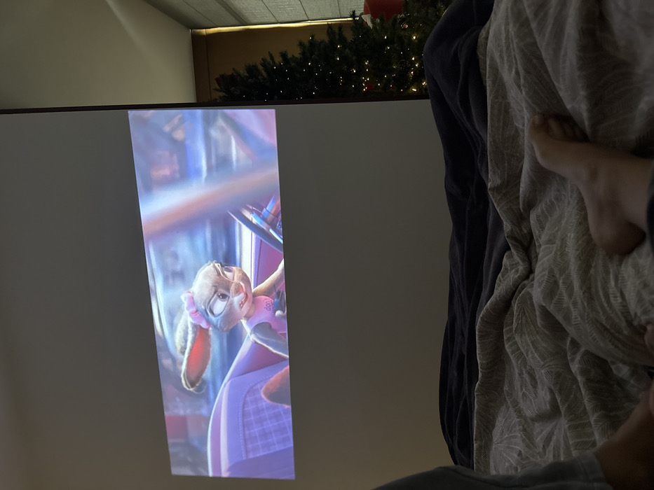 The image captures an indoor scene in Warsaw, Poland, featuring a projected animation on a light-colored wall. The projection displays the animated character Judy Hopps from Disney's Zootopia, depicted as a rabbit wearing a pink top with "don" partially visible, a pink hair bow, and seated in the driver's seat of a pink car, exhibiting an assertive facial expression. In the foreground, the lower legs and feet of an individual are visible, resting on a bed covered with a light-colored, patterned duvet and a dark grey or black blanket. To the right of the projection, a dark green Christmas tree adorned with white lights is partially visible, indicating a decorative element within the room. The lighting in the room is dim, suggesting an intentional setting for viewing the projection.