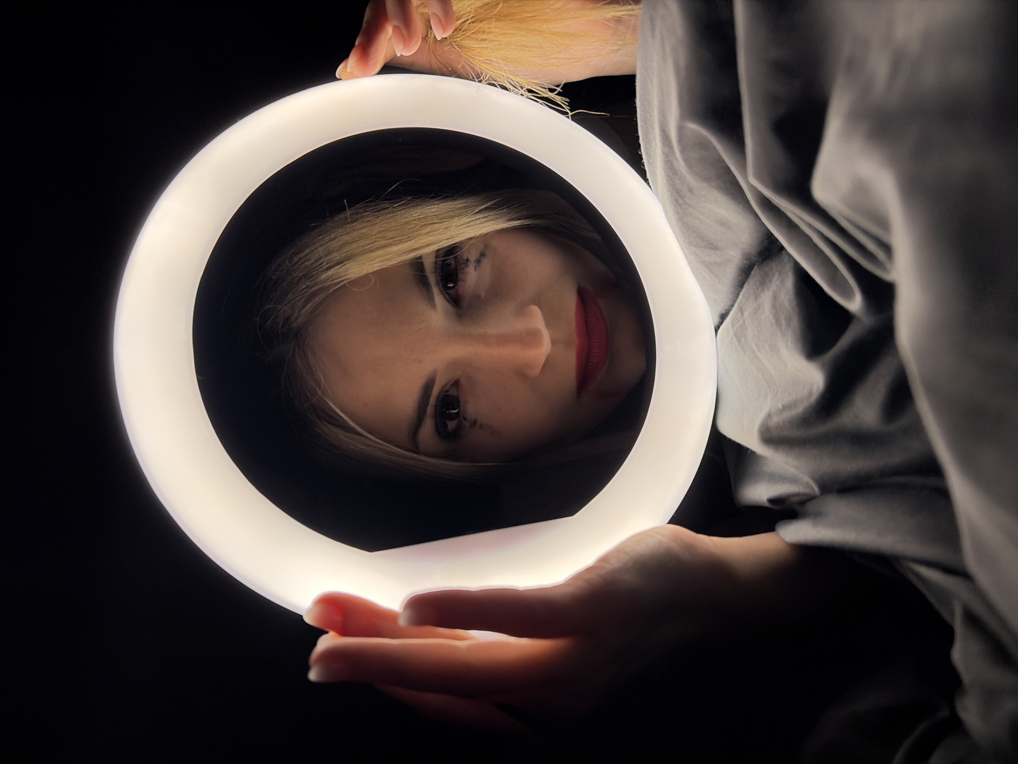 A fair-skinned woman with blonde hair is seen through a ring light, the device itself held by her hands. She is wearing red lipstick and has dark mascara. The background is black. Part of a gray garment is visible on the right. The location is not evident from the image.