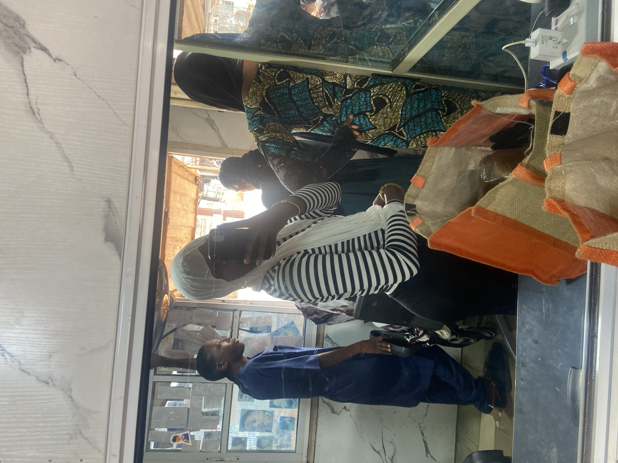 The image shows a reflection in a mirror, likely inside a business establishment, possibly a beauty salon or tailoring shop, in Okengwe - Obehira, Nigeria. Inside the shop, there are at least three people. One person is wearing a striped shirt and a head covering, using a mobile phone, possibly taking a picture of the mirror. Another person is dressed in a blue outfit and appears to be standing or sitting near a window with pictures or posters displayed on it. A third individual is wearing a patterned dress. There is a tote bag hanging close to the mirror.