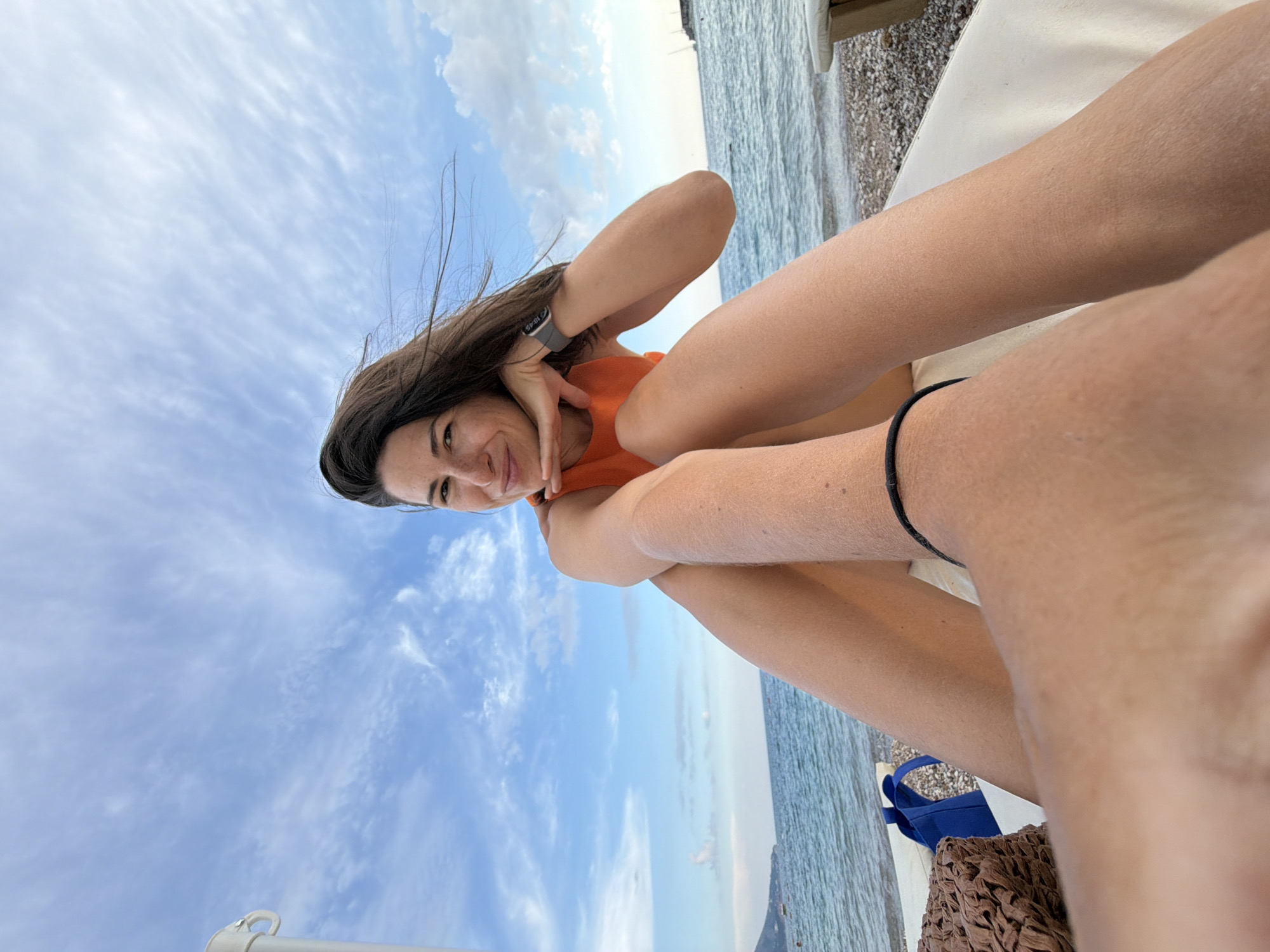 A woman is pictured from a first-person perspective, reclined on a beach lounger on a pebbly beach with the Adriatic Sea and a cloudy sky in the background. She is wearing an orange top and a watch on her left wrist. The image appears to have been taken in Budva, Montenegro.