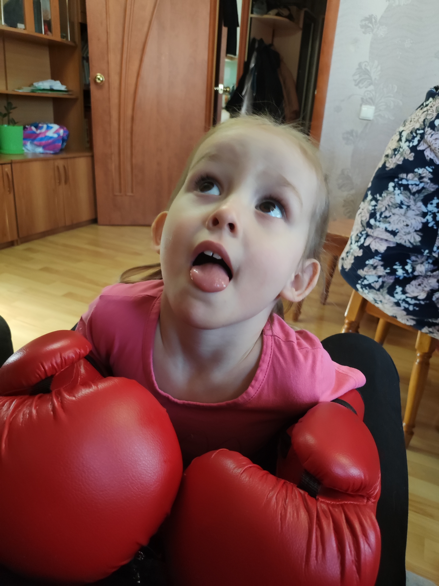 A young child wearing a pink shirt is seated with two large red boxing gloves resting on their lap. The child has blonde hair pulled back and is sticking their tongue out while looking upwards. The background includes a wooden door, a cabinet, wallpaper with a floral pattern, and a wooden floor. The location appears to be indoors, possibly a home. There are no indications about being in Nizhnyaya Gar', Russia.